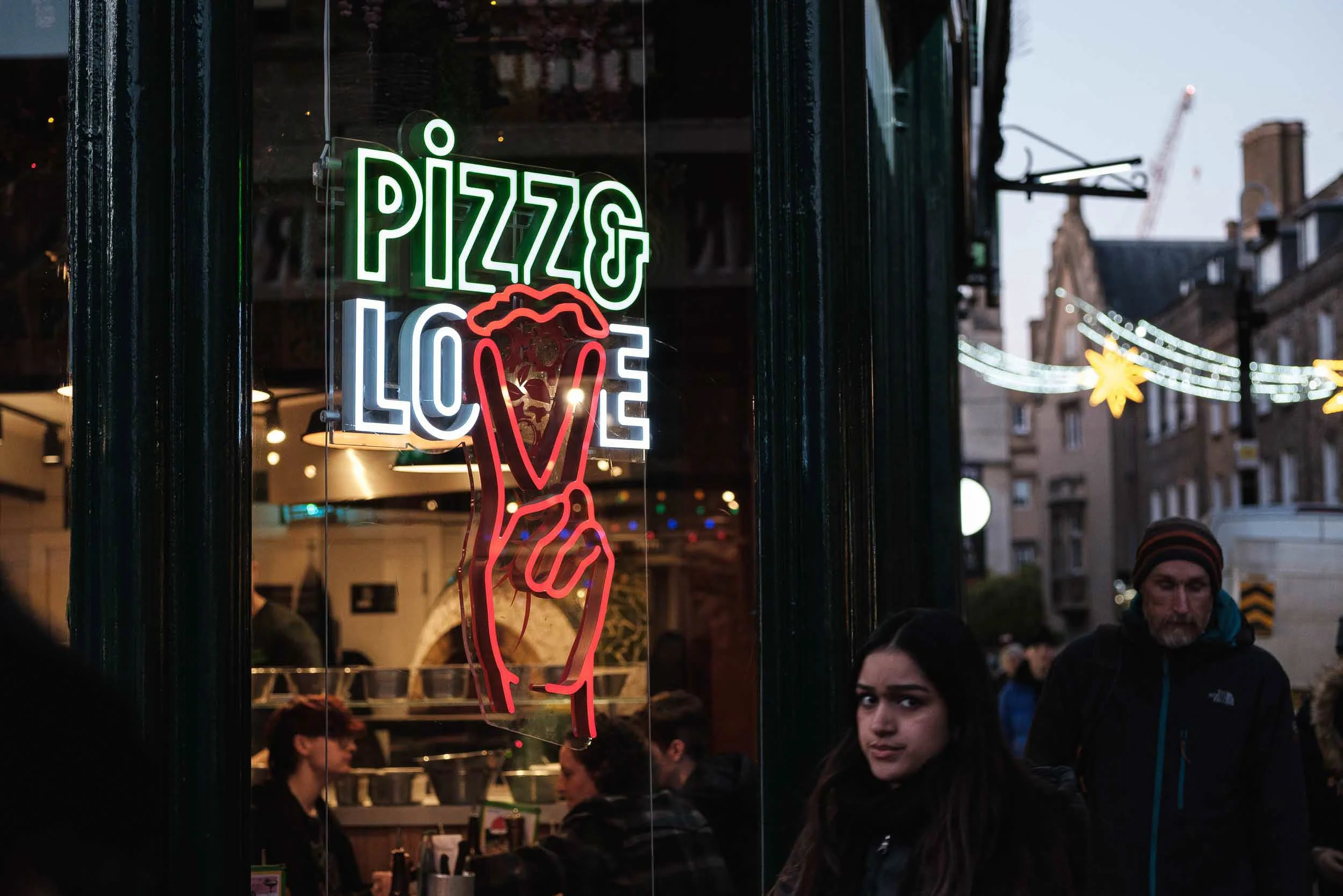 Neon sign on restaurant window reading 'Pizza Love' with a hand holding a slice of pizza, and people dining inside and outside.