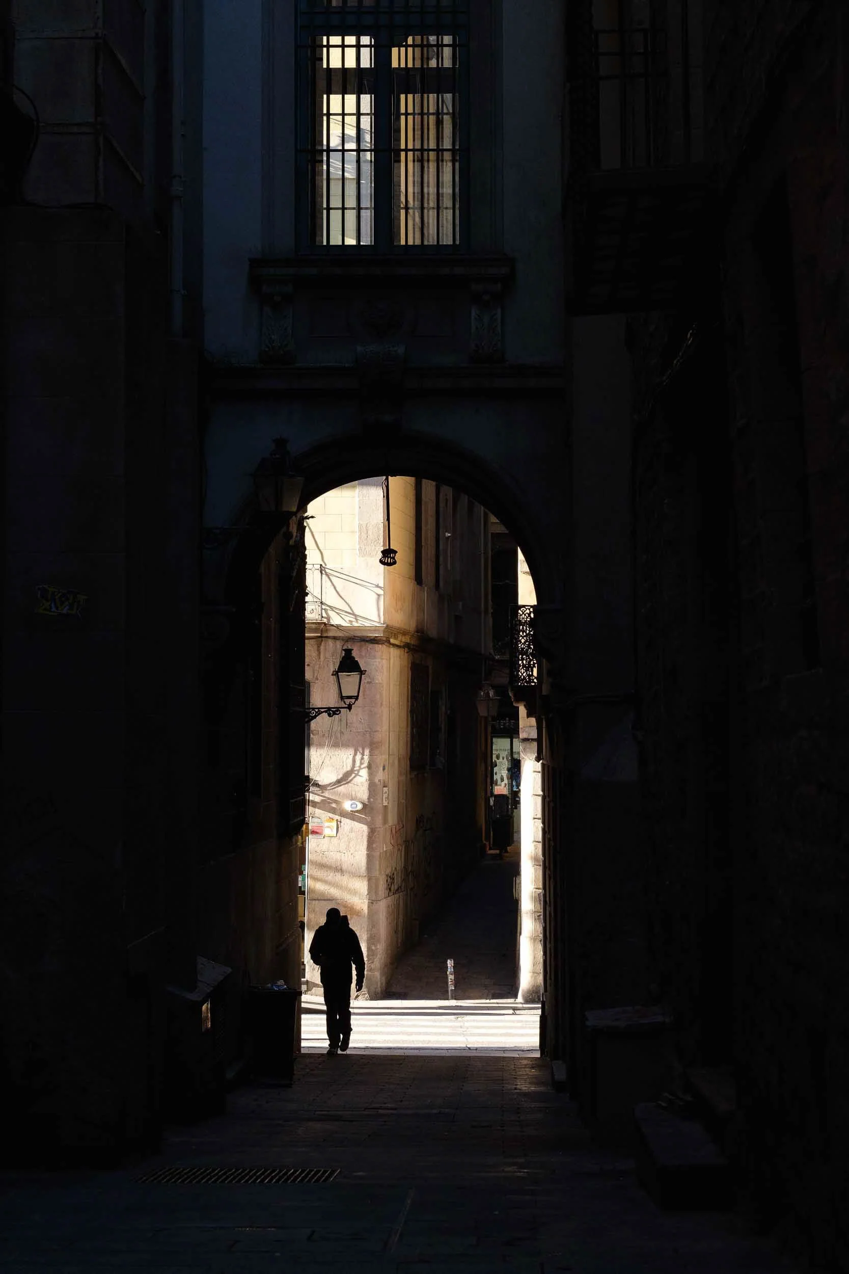 Dark alleyway with archway opening to a sunlit street, silhouetted person walking towards the daylight, buildings with balconies and lanterns in the background.