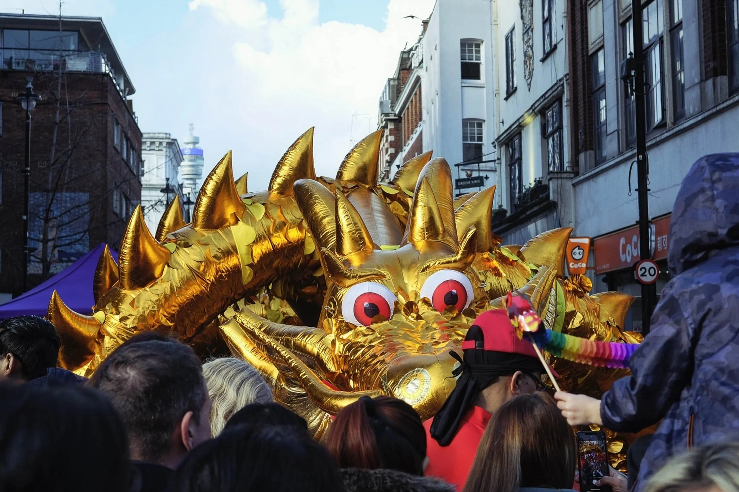 A golden dragon with big red eyes looks at people in a crowd with a child holding a small dragon figure in Chinatown London and BT tower in the distance