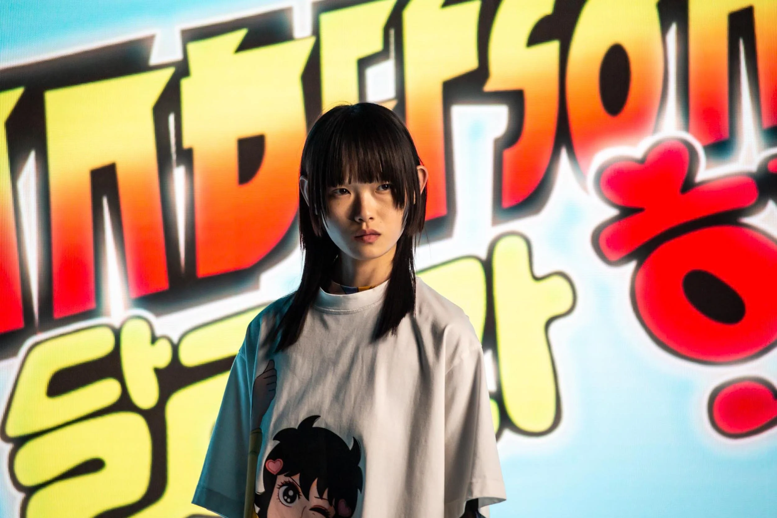 A woman wearing a tshirt with anime character on stands in front of Japanese neon grafitit on a LED screen onset at a fashion shoot