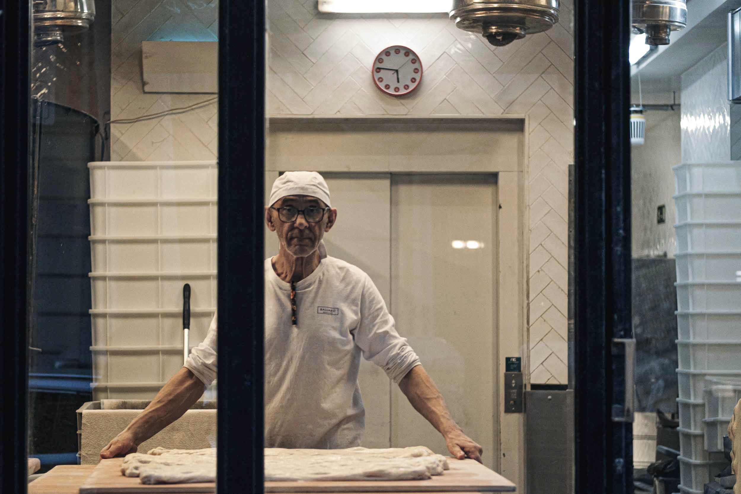A chef in a white uniform and cap is working behind a glass window at a bakery or pizzeria, preparing dough on a wooden table.