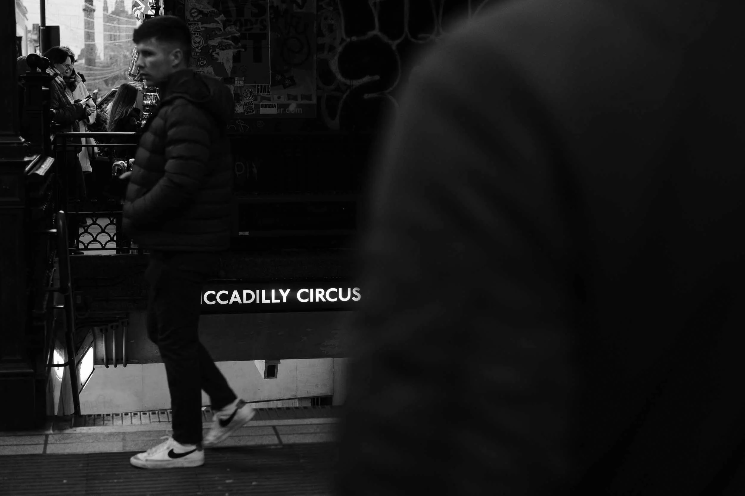 Layered black and white image outside Piccadilly Circus station entrance busy with people and glowing station sign
