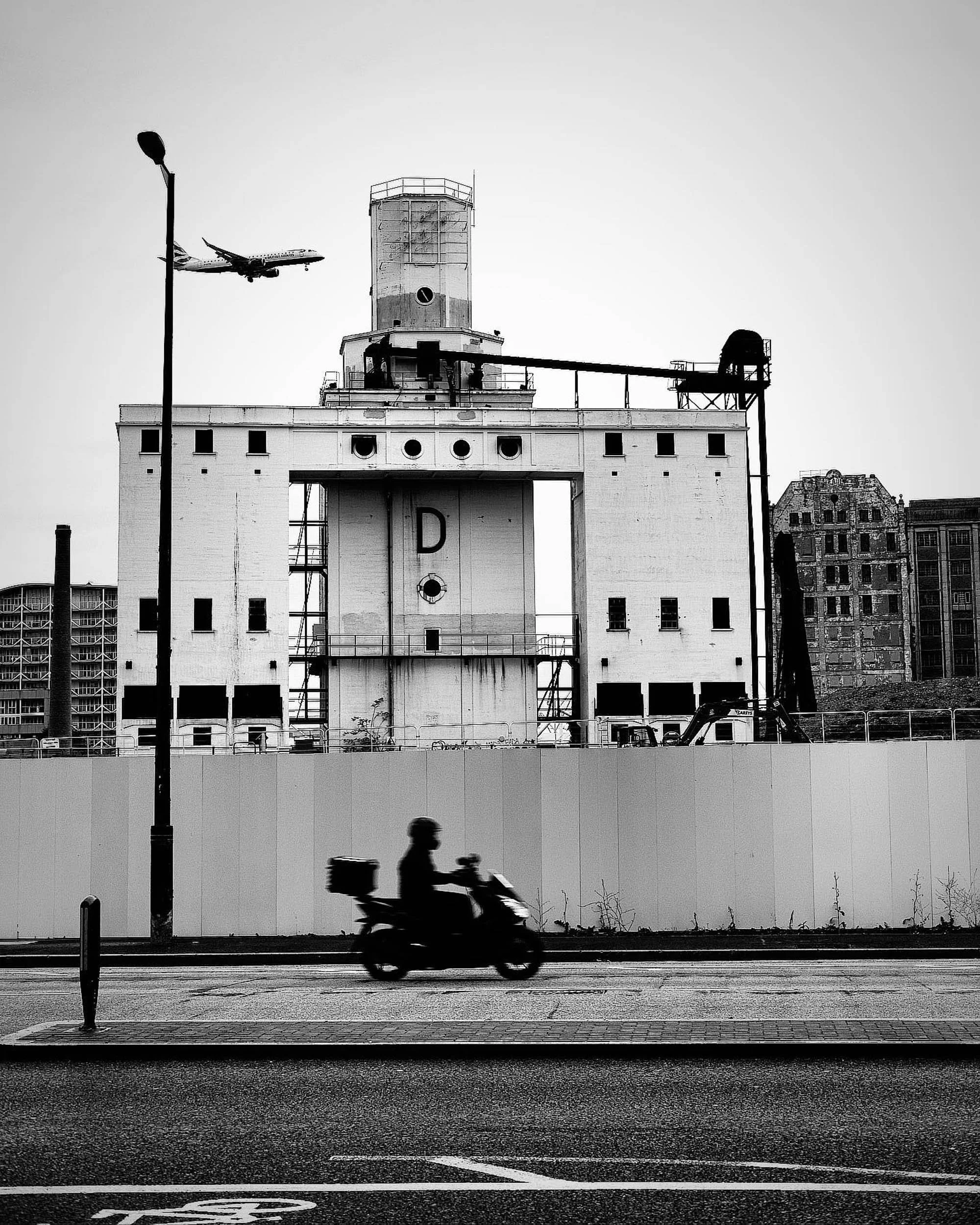 Strong black and white image in east london development with a moment in time frozen as a bike passes by with a plane in the background