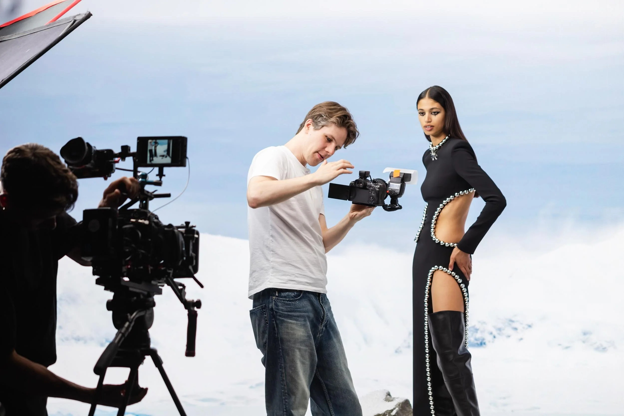 A videographer checks settings as a woman poses in a black revealing dress set against a white snowy background created on large LED screen for fashion brand David Koma
