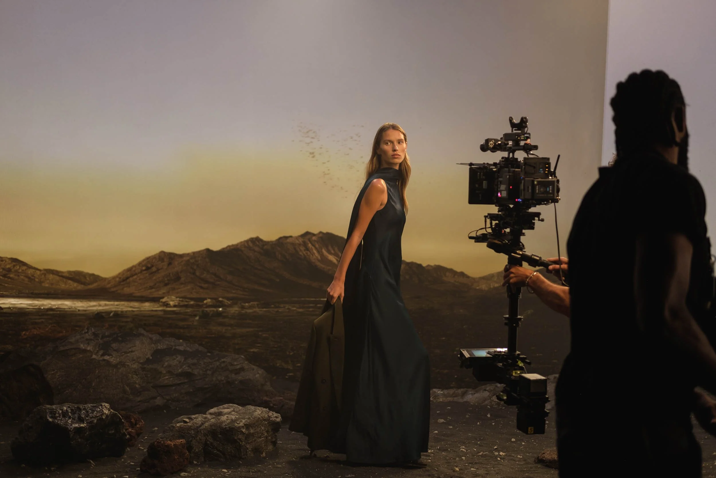 A woman with long, wavy hair standing in front of a virtual desert landscape backdrop shown on large LED Screen during a film shoot. She is wearing a black dress and carrying a tan handbag.