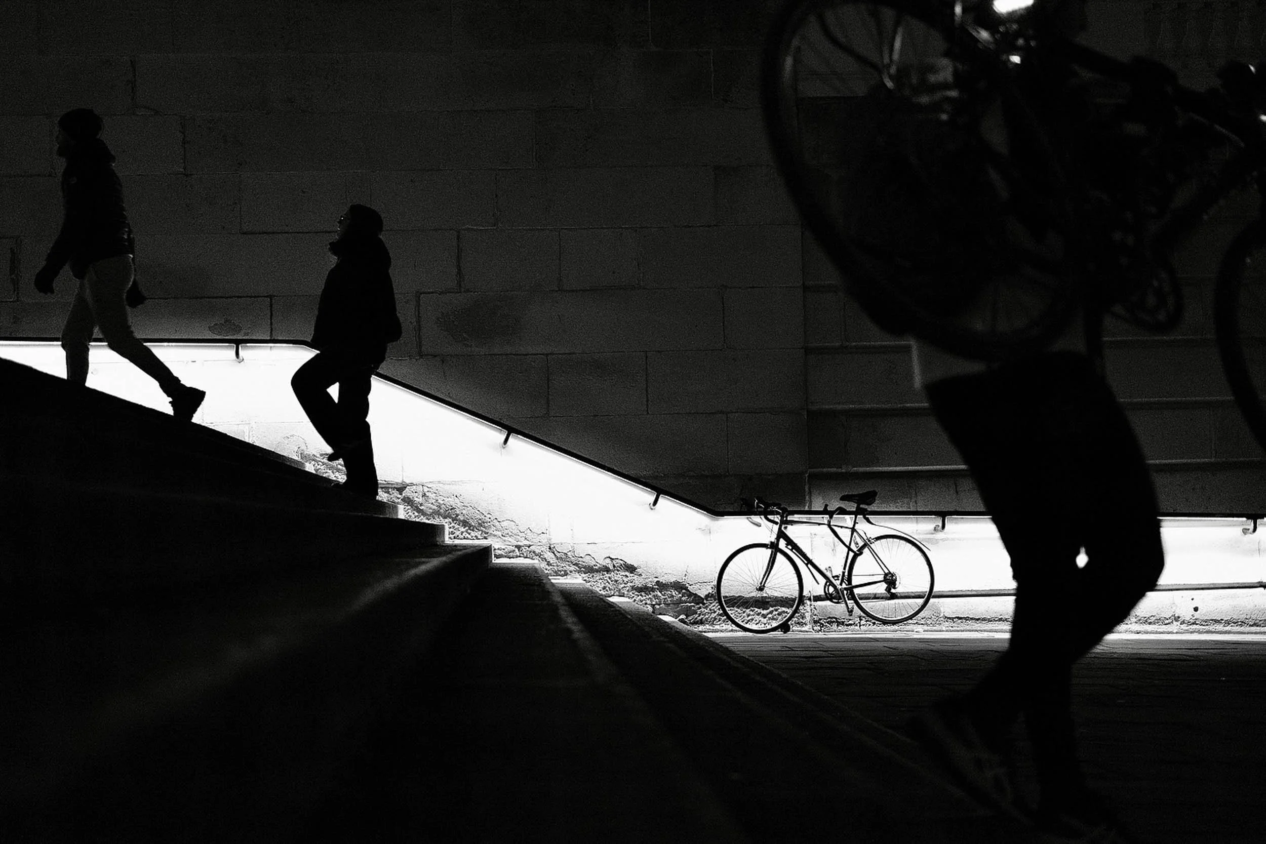 silhouettes of a man carrying a bike down steps in black and white with a bike in the background creating geometry and layers to this night scene in London