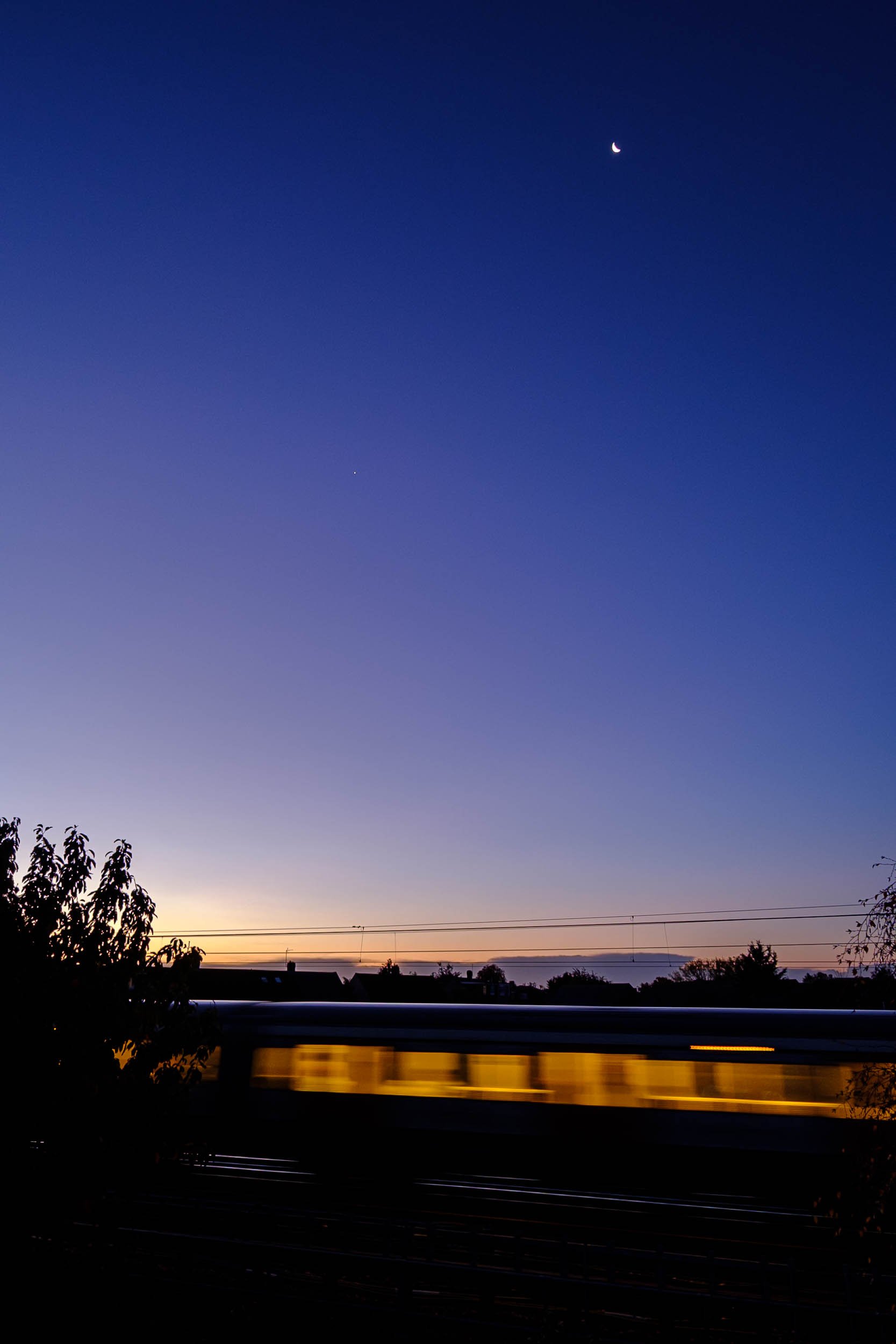 Dawn colours of blue purple and yellow in Essex as the warm light from a passing underground train gives motion blur with the moon visible