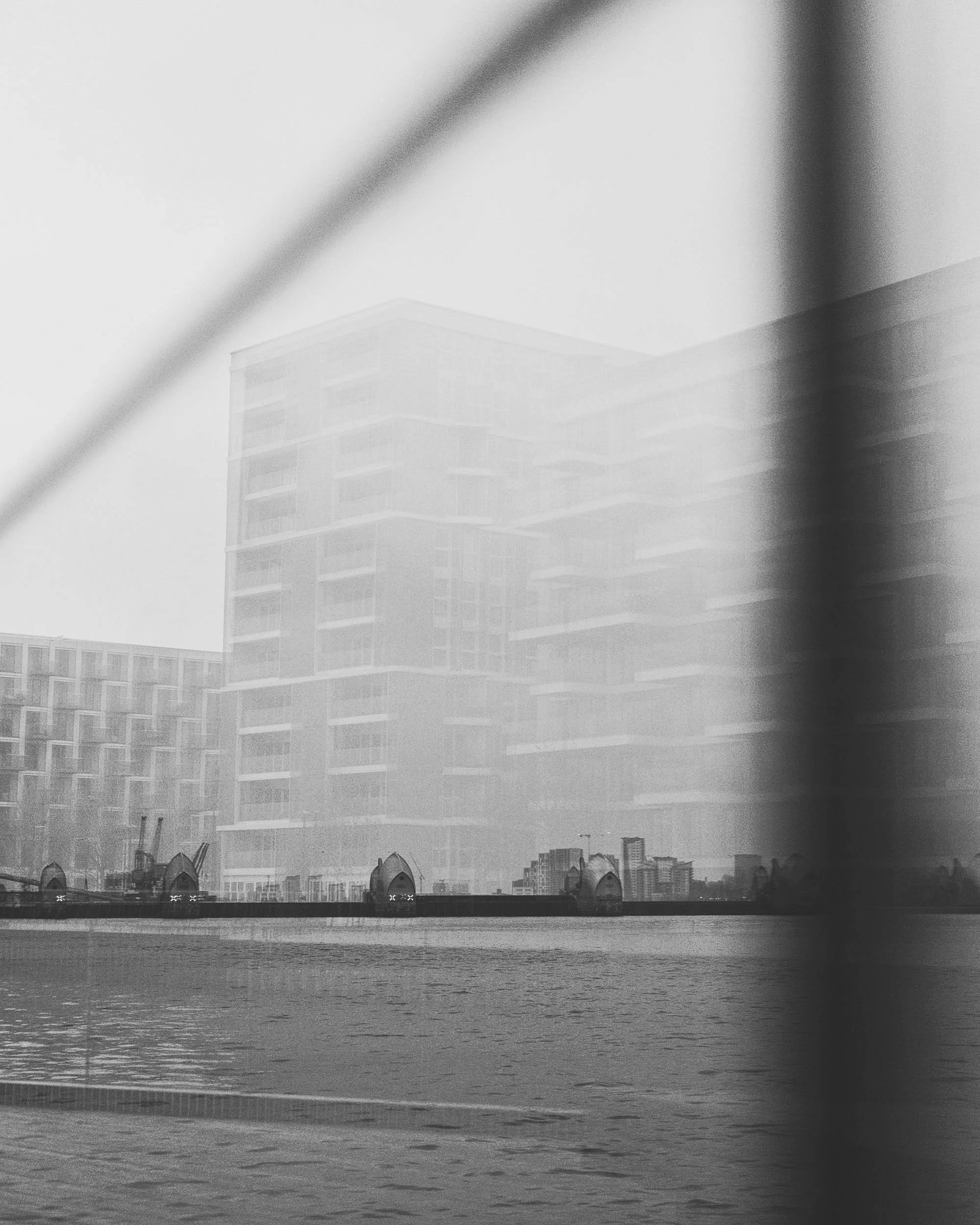 A black and white photo of a city skyline viewed across a body of water, with modern buildings in a reflection that adds layers to the scene, showing the Thames Barrier in East London.