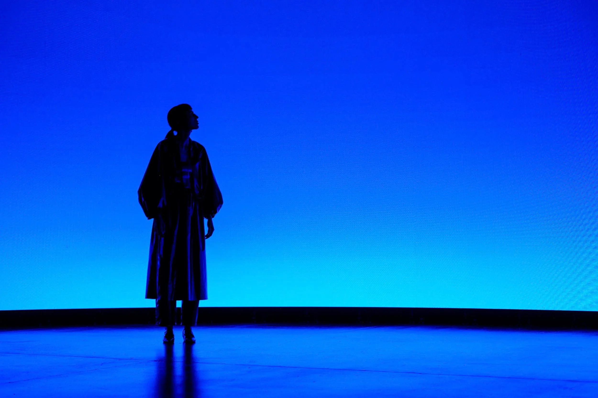 Woman standing in shiny dark raincoat against a blue gradient background from a large LED screen as a virtual production shoot for drinks brand Tanqueray Gin