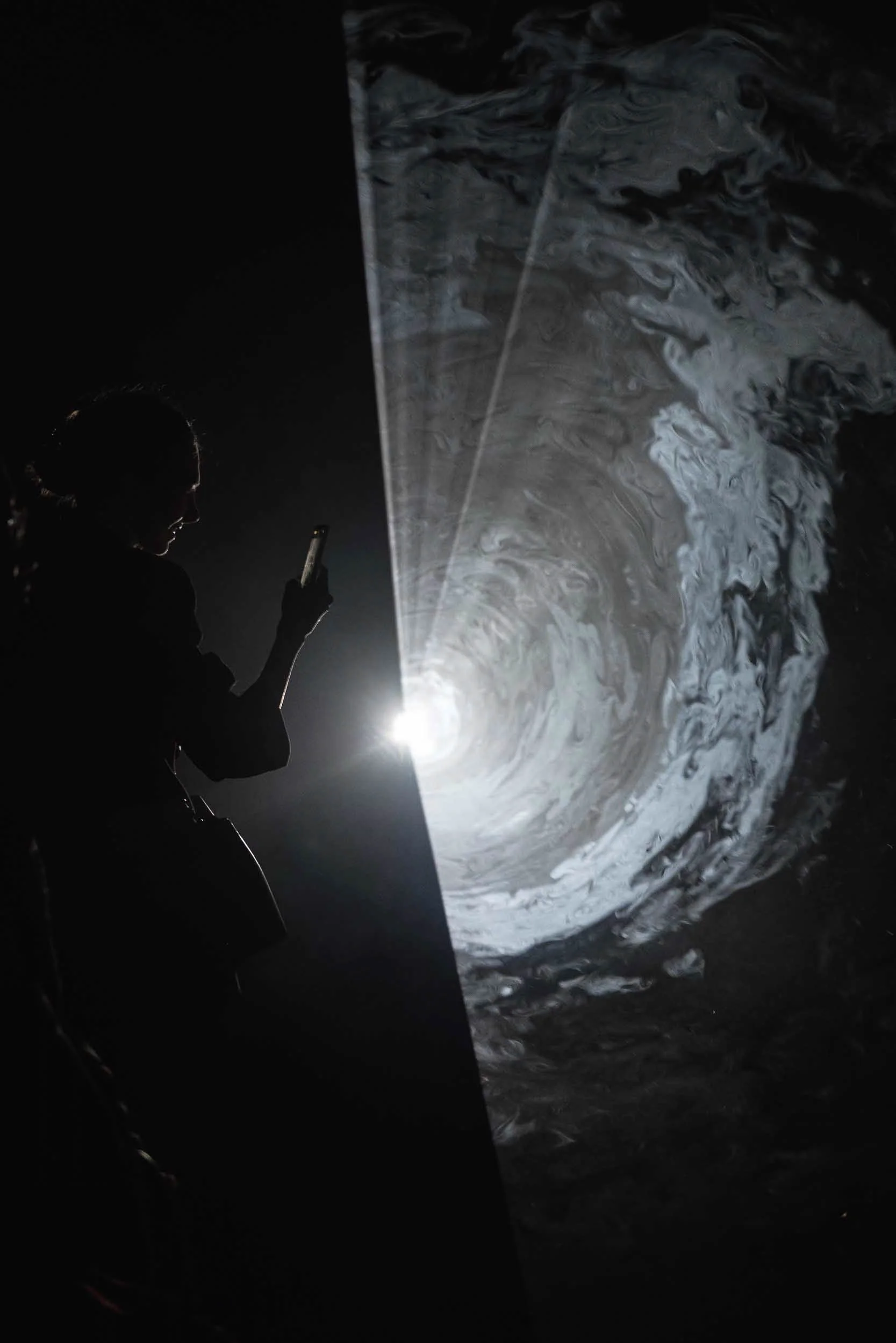 A woman takes a picture with a smartphone of a intense beam of light at a interactive art show at Tate Modern in London set against a dark black background