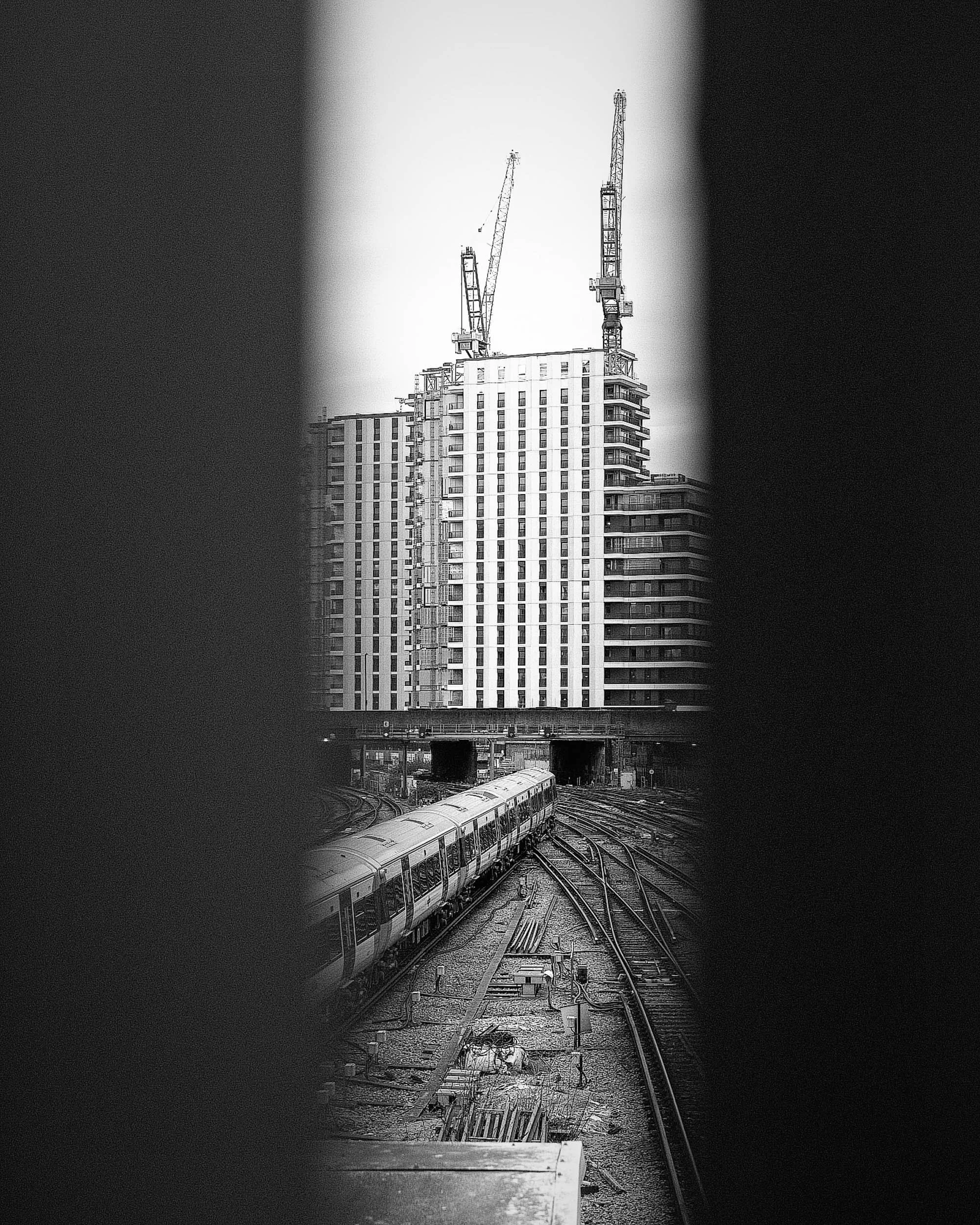 Black and white photo of a high-rise building under construction with cranes on top, rail tracks in the foreground, and a train passing by underneath within a thin piece of the frame with large portions obscured adding to the vertical composition.