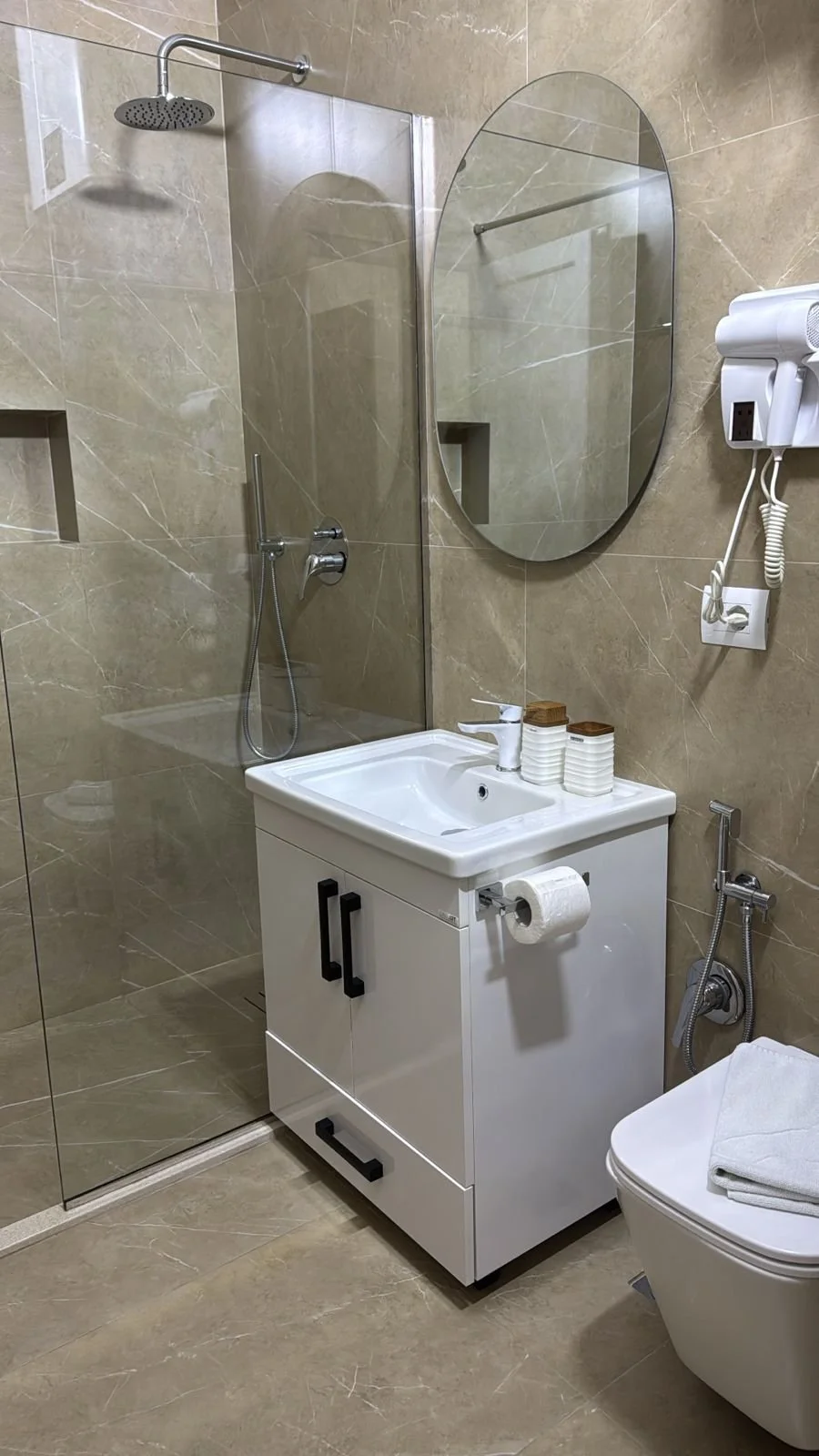 Bagno moderno con pareti e pavimenti in beige marmo, doccia con parete di vetro, lavabo bianco con mobile sottostante, specchio rotondo, asciugacapelli wall-mounted, WC con carta igienica.