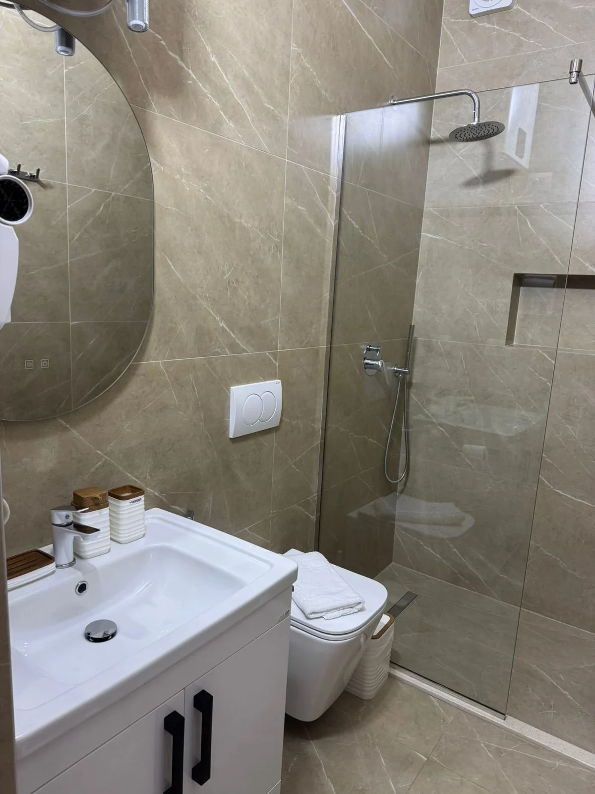 Bagno con pareti in piastrelle beige, lavandino bianco con due contenitori di sapone e una tazza, soffione doccia rotondo sopra una parete divisoria in vetro, e un WC con bidet. Sono presenti anche un armadietto con maniglie nere, e alcuni asciugaman