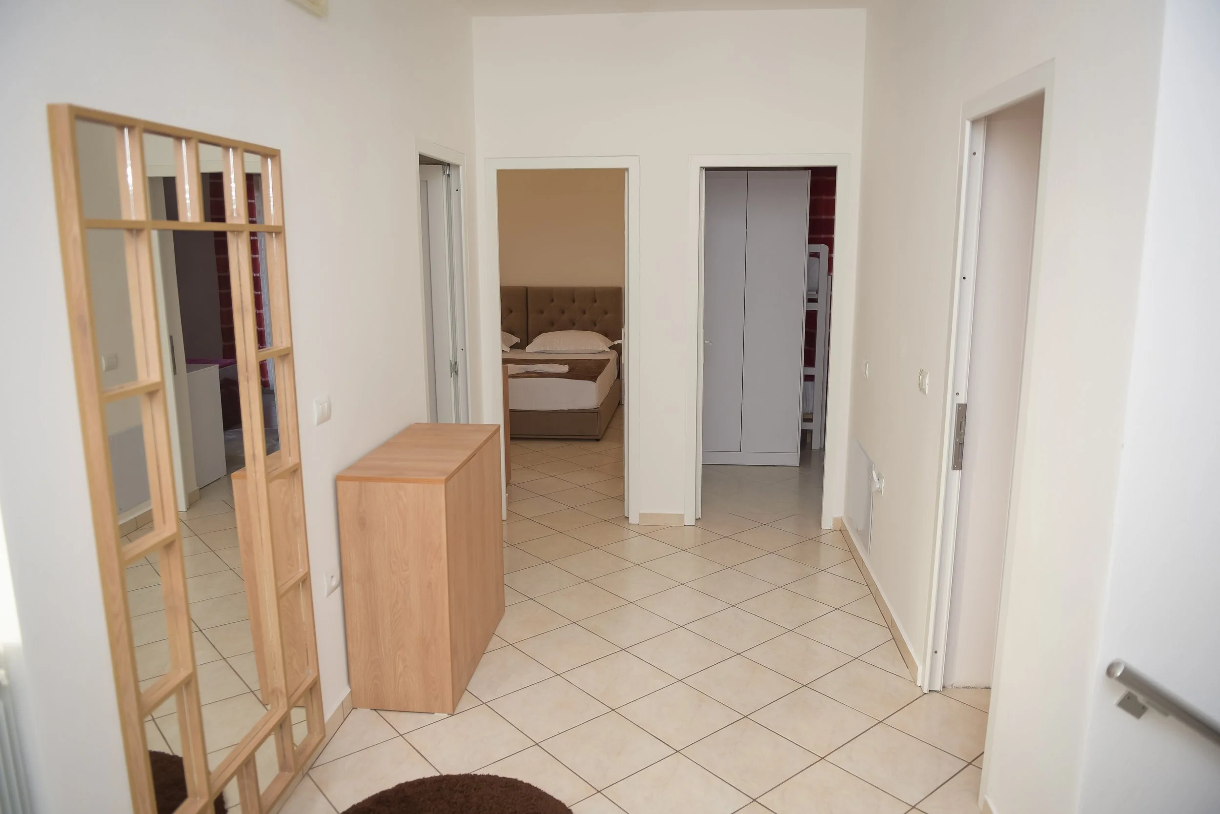Ingresso di un appartamento con vista su varie stanze, pavimento piastrellato, pareti bianche, porta bianca aperta su una camera con letto e cuscini, armadio grigio e porta di una lavatrice, divisorio in legno decorativo, tappeto rotondo marrone.