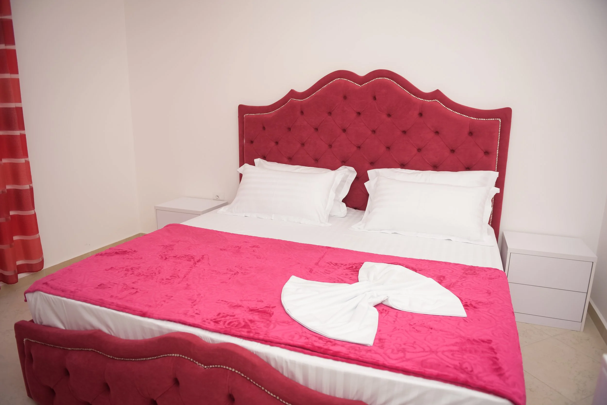 Letto matrimoniale con testiera rossa in tessuto, decorata con bottoni, con lenzuola bianche e un copriletto rosa, e due cuscini bianchi. Ai lati, due comodini bianchi. A sinistra, una tenda rossa con strisce, e una parete bianca