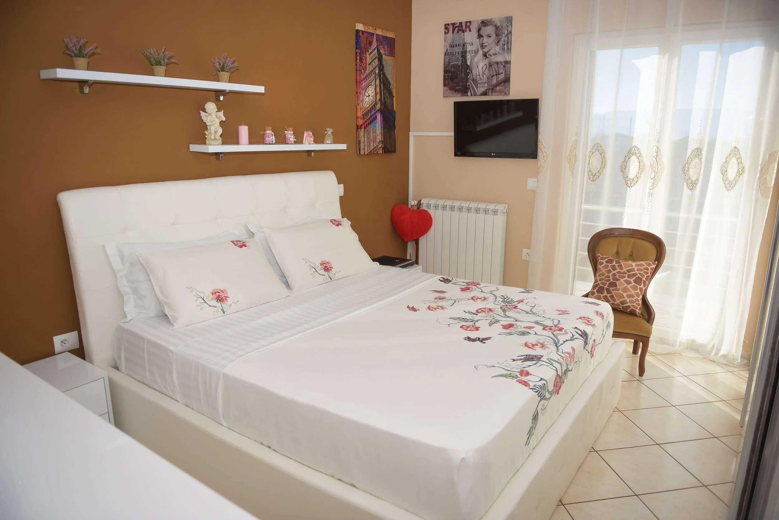 Camera da letto con letto matrimoniale con biancheria a righe e fiori, pareti color cioccolato, scaffali con decorazioni, quadro di città e schermo TV, sotto una finestra con tende chiare, sedia con cuscino a motivo giraffe e decorazioni come statuet