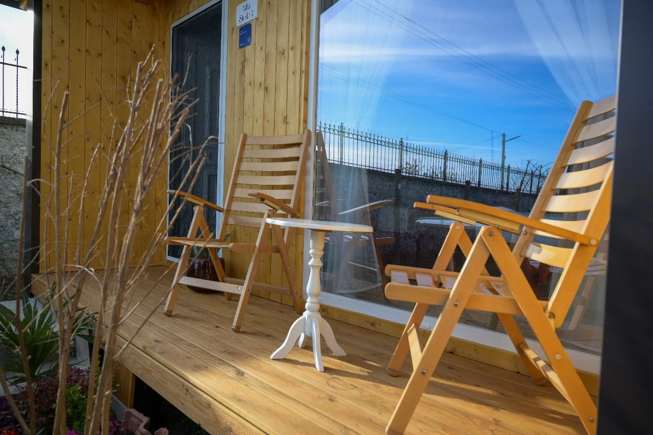 Balcone di legno con due sedie pieghevoli e un piccolo tavolino bianco, davanti a una finestra con tende trasparenti, con un cielo azzurro e fili dell'elettricità visibili.