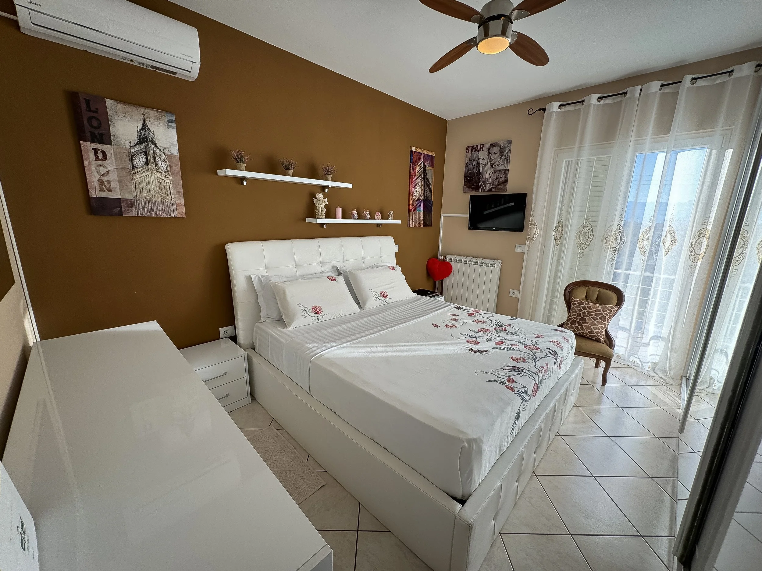 Camera che mostra una stanza da letto con parete marrone, quadri, una televisione, un ventilatore a soffitto, un condizionatore, tende bianche e un letto con lenzuola a fiori.
