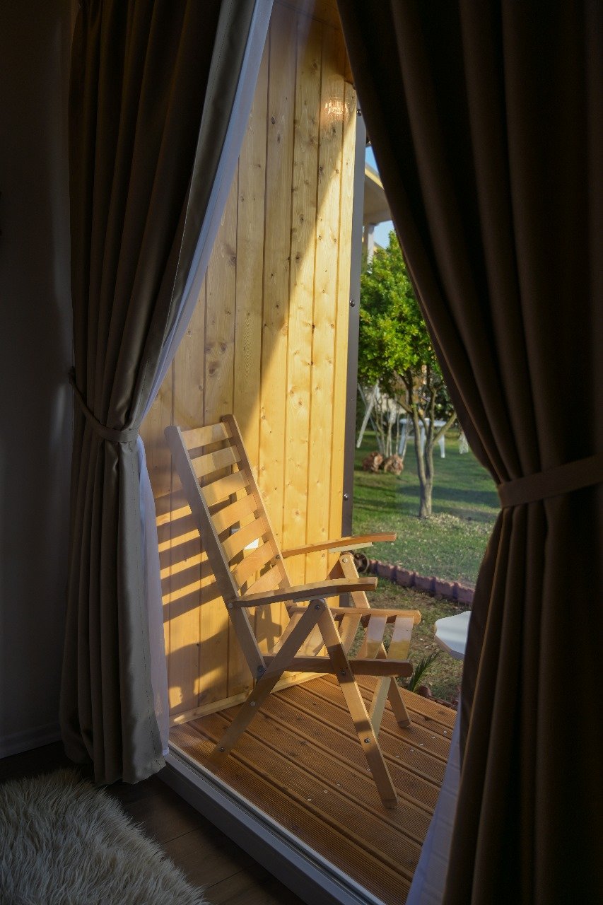 Sedia di legno su un balcone di legno, visto attraverso una porta scorrevole con tende marroni e una parete di legno di sfondo, con un giardino con alberi e prato all'aperto.