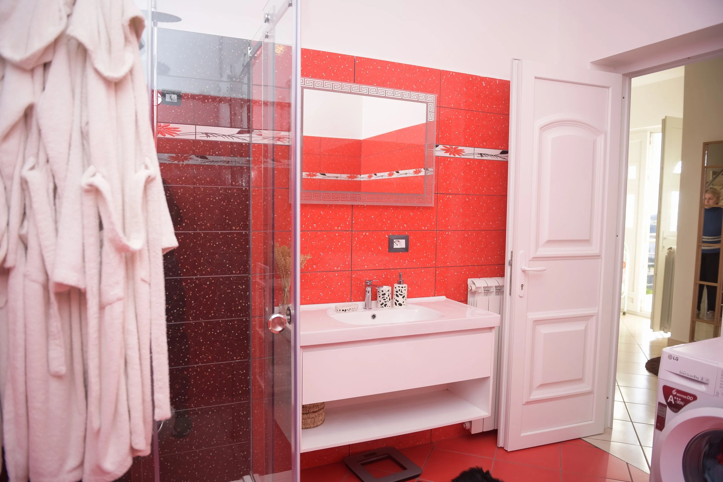 Bagno con pareti rosse e specchio sopra il lavabo, porta bianca, asciugamani rosa, lavatrice visibile alla destra, tappeto nero sul pavimento, e un individuo in sfondo.