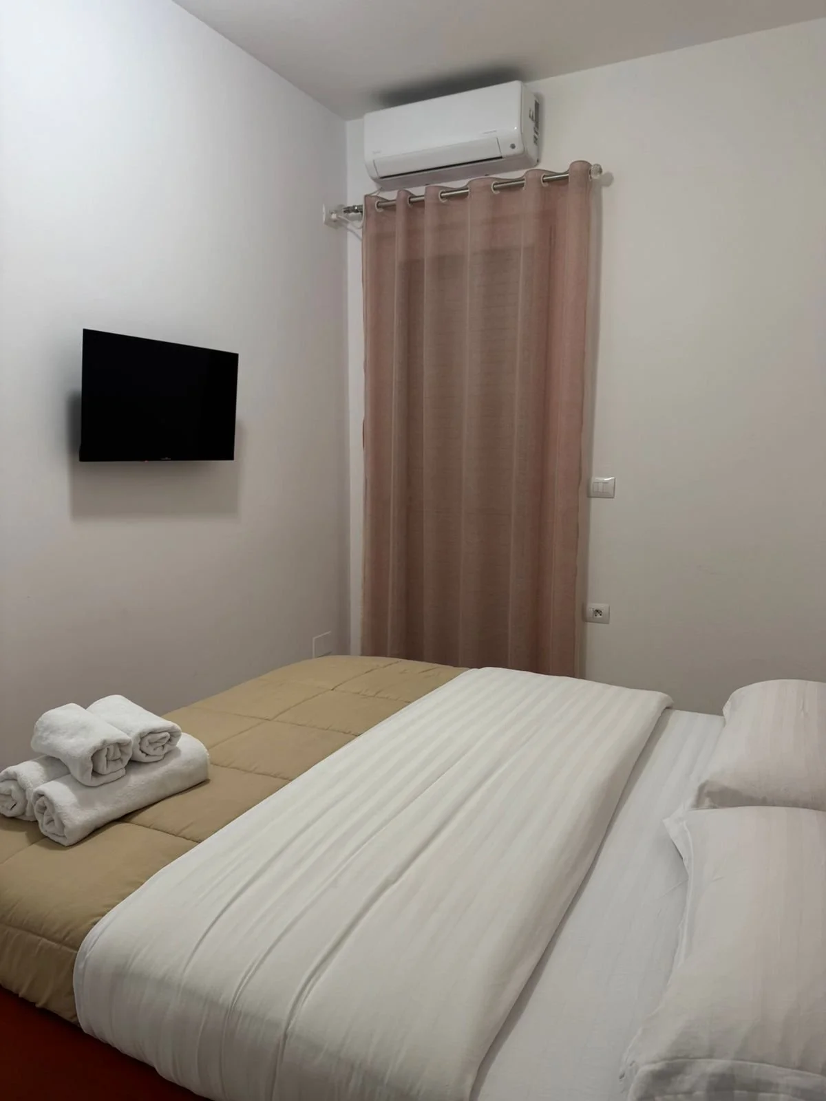 Camera da letto con letto matrimoniale, asciugamani arrotolati sul letto, televisore a schermo piatto sulla parete, condizionatore d'aria, tende marroni, pareti bianche.