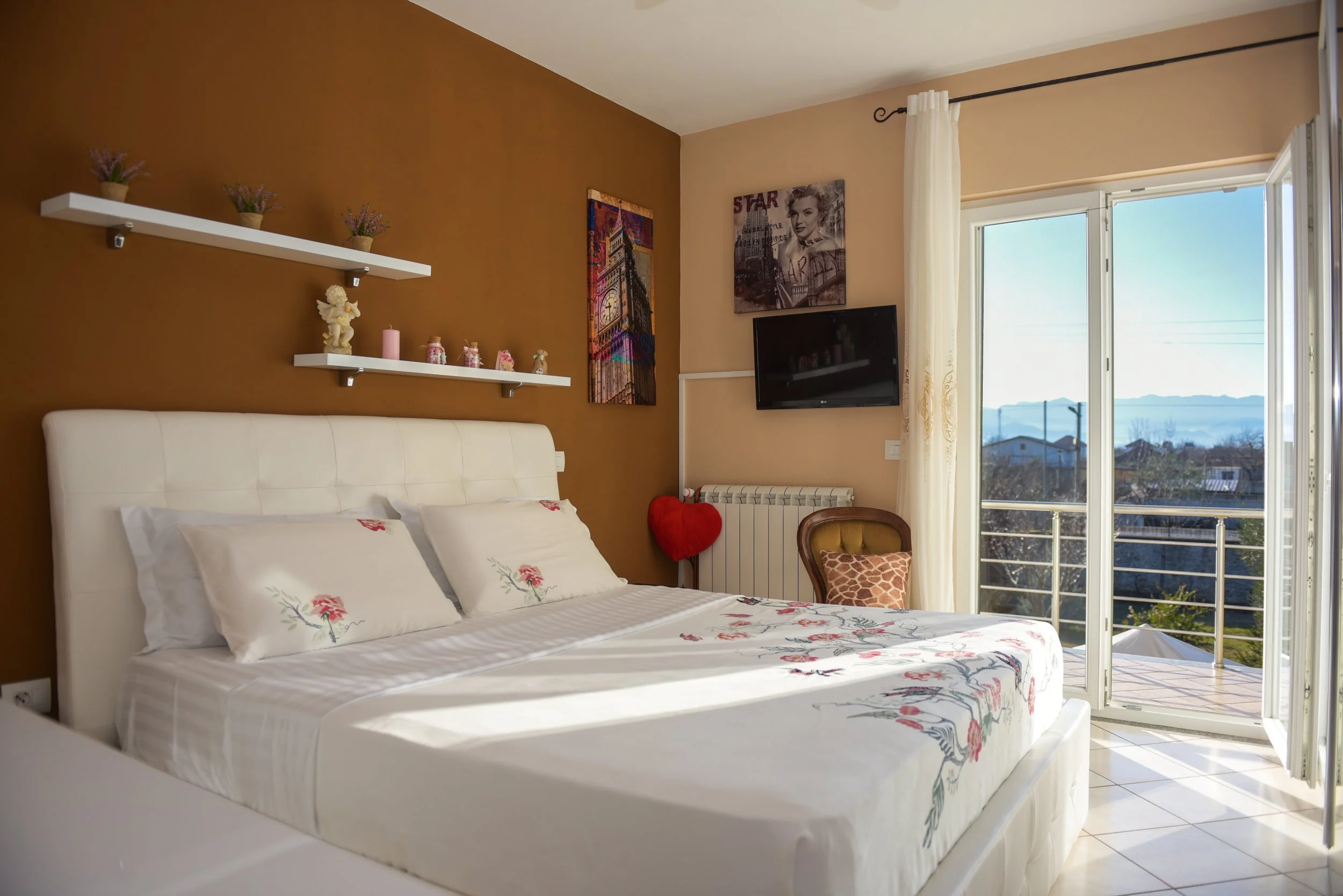 Camera da letto con letto matrimoniale, coperte e cuscini con ricami floreali, pareti color cioccolato e beige, mensole con vasi di lavanderia e decorazioni, televisore, dipinto di Marylin Monroe, finestra con tendina e vista panoramica di colline e 
