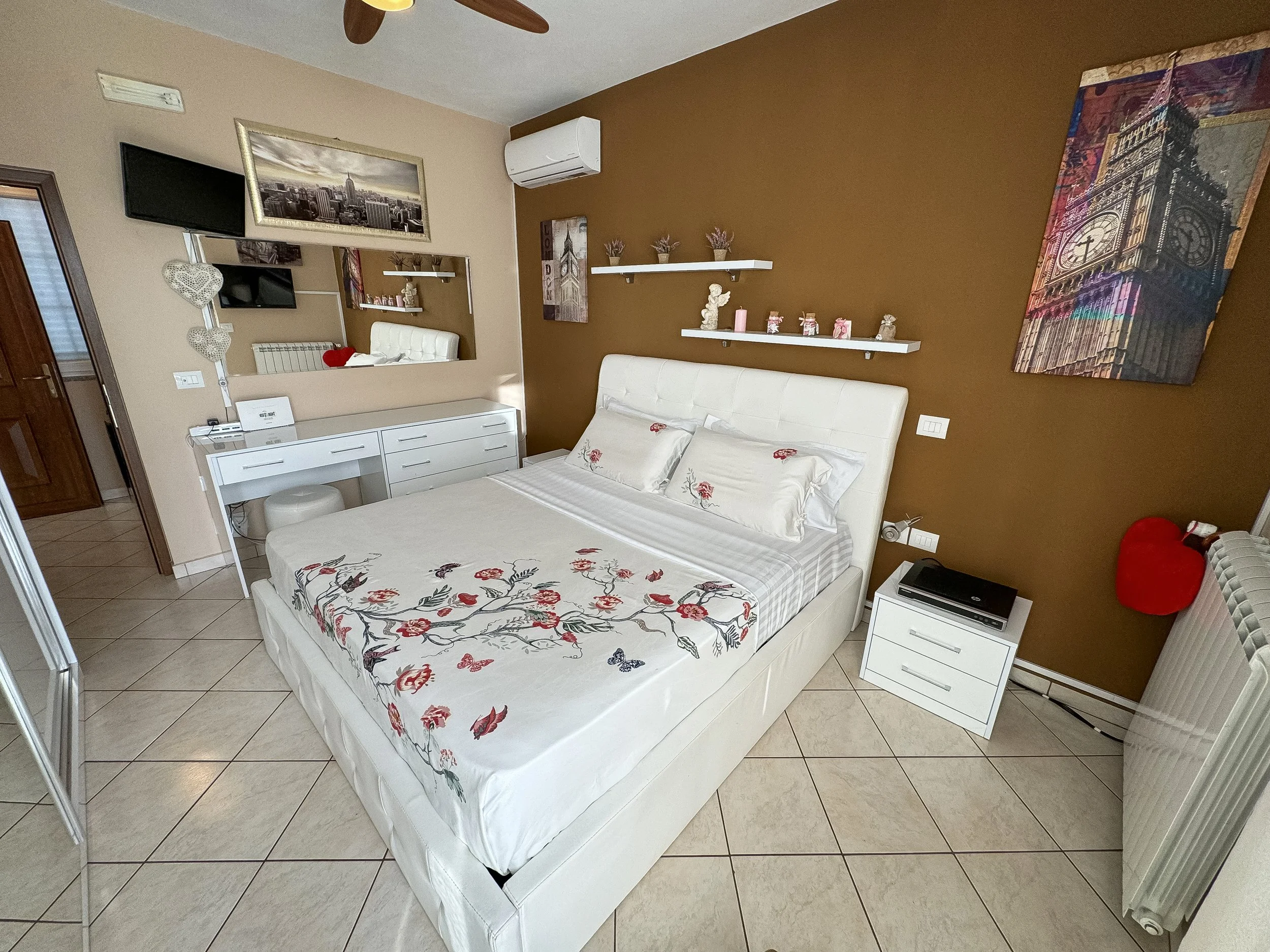 Camera da letto con letto con biancheria bianca decorata con fiori e farfalle rosse e blu, parete marrone con scaffali bianchi e decorazioni, quadro di Big Ben e skyline di New York, televisore e aria condizionata, mobili bianchi, vari oggetti decora
