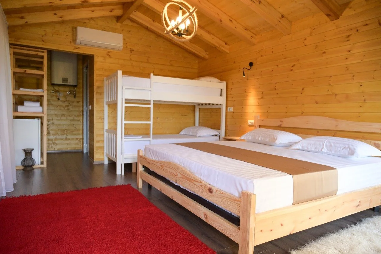 Interno di una camera in stile rustico con pareti di legno, letto matrimoniale con cuscini bianchi, letto a castello in lato, tappeto rosso e soffitto a volta in legno.