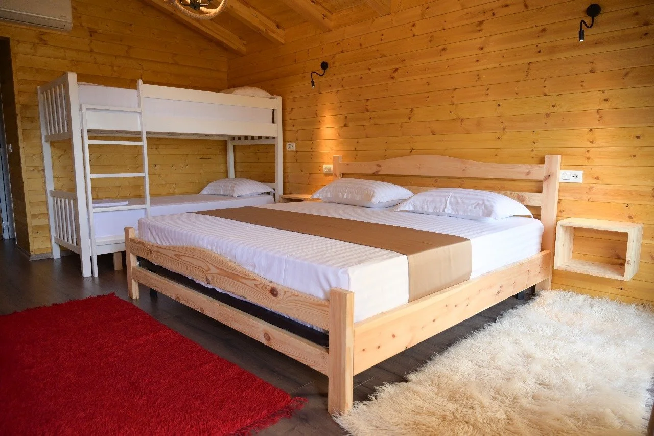 Camera in stile rustico con pareti di legno, letto matrimoniale in legno con cuscini e lenzuola bianche, letto a castello bianco con lenzuola e cuscini, tappeti rossi e beige, luci a parete di fronte al letto.