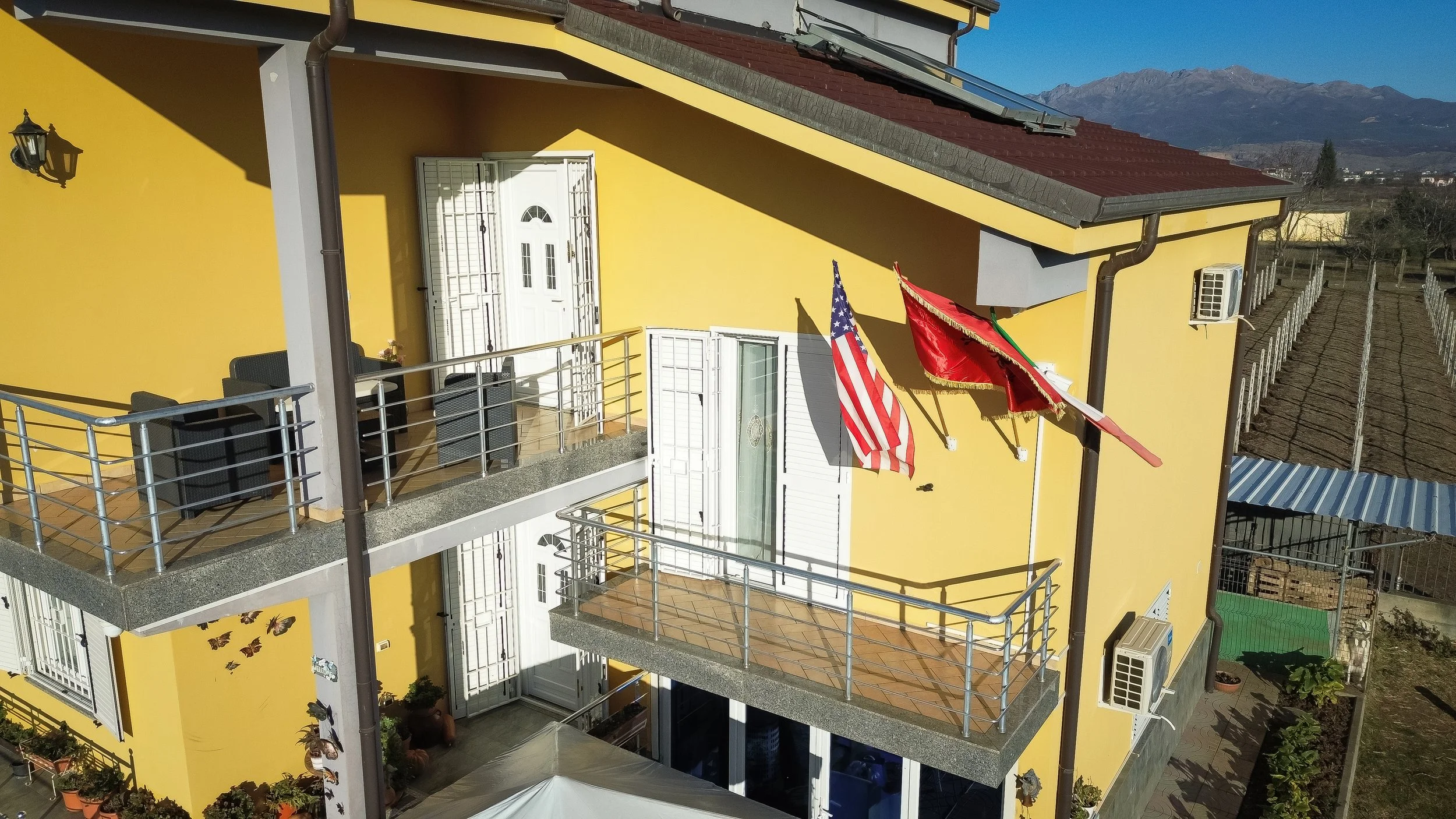 Edificio di civile abitazione color giallo con balconi, bandiere americana e italiana, piante sul balcone e in terrazza, aria condizionata. Sullo sfondo si vedono montagne e un cielo blu.