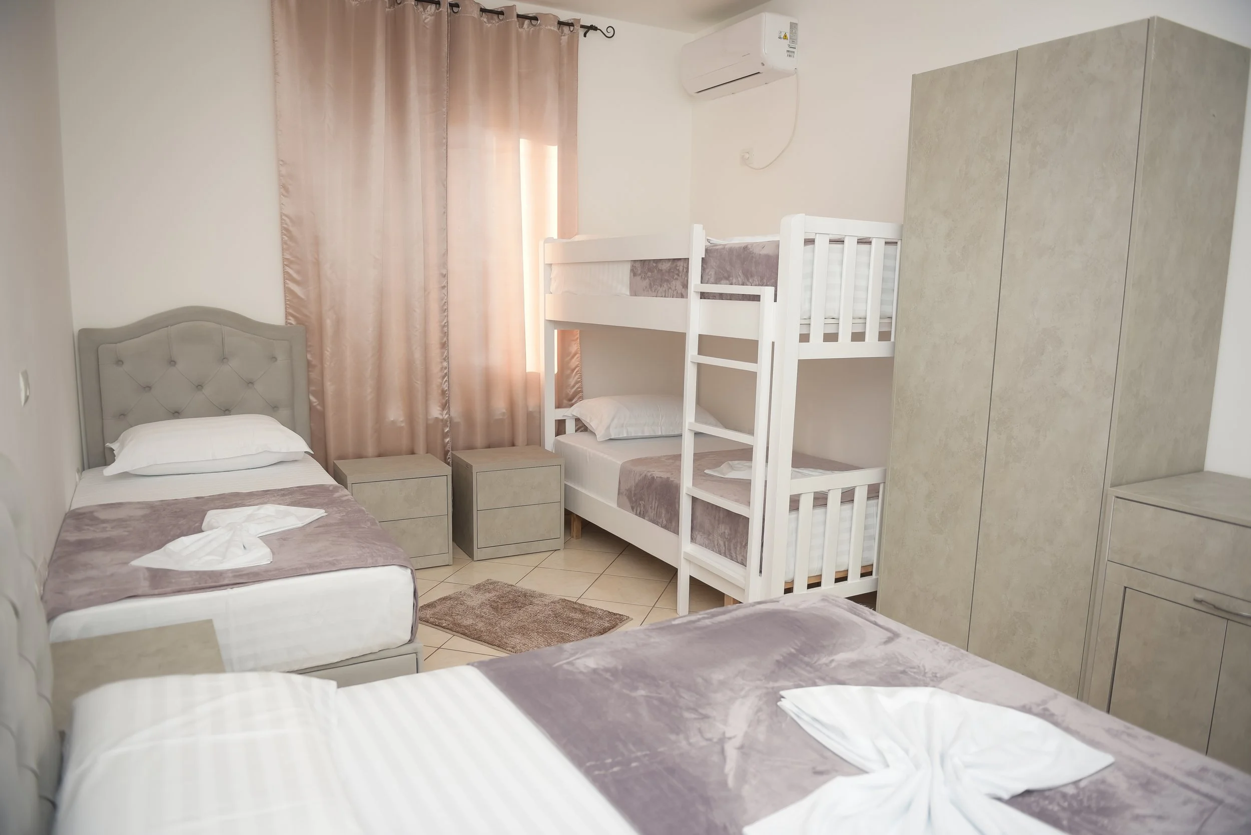 Camera con tre letti singoli e un letto a castello, luce naturale dalle tende, aria condizionata, armadio e tappeto beige