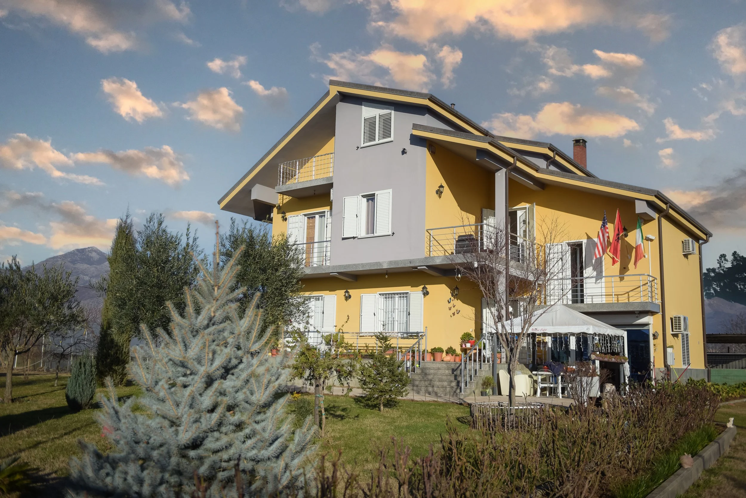 Una casa a più piani color giallo e grigio con balconi e bandiere davanti, circondata da un giardino con alberi e piante, sullo sfondo montagne e un cielo con alcune nuvole.