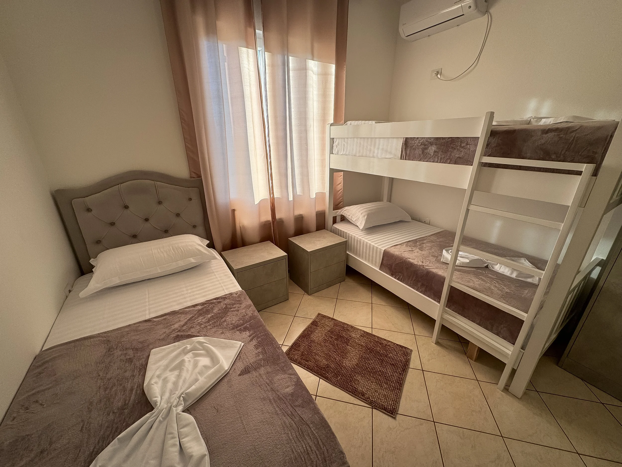 Camera rivolta verso una stanza con due letti singoli, un armadietto di plastica tra i due letti, e un secondo letto a castello. Le pareti sono di colore chiaro, con una finestra con tende di colore rosa e beige. Sopra il letto a castello c'è un cond
