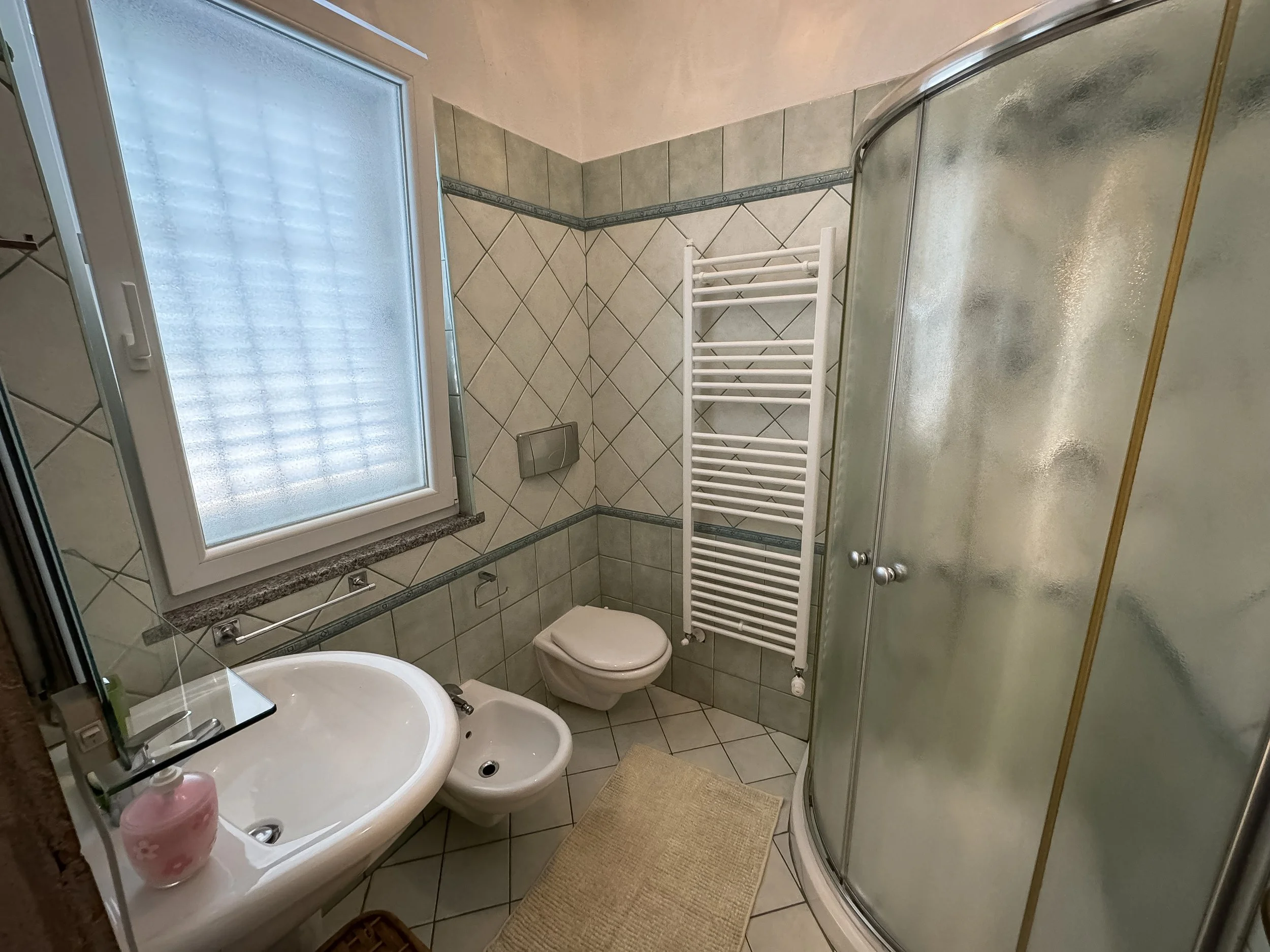 Una vista di un bagno con pareti rivestite di piastrelle beige e decorative. Include una finestra con vetro satinato, uno specchio sopra un lavandino bianco, un bidet, un WC, un termosifone bianco e una cabina doccia con pareti in vetro smerigliato. 