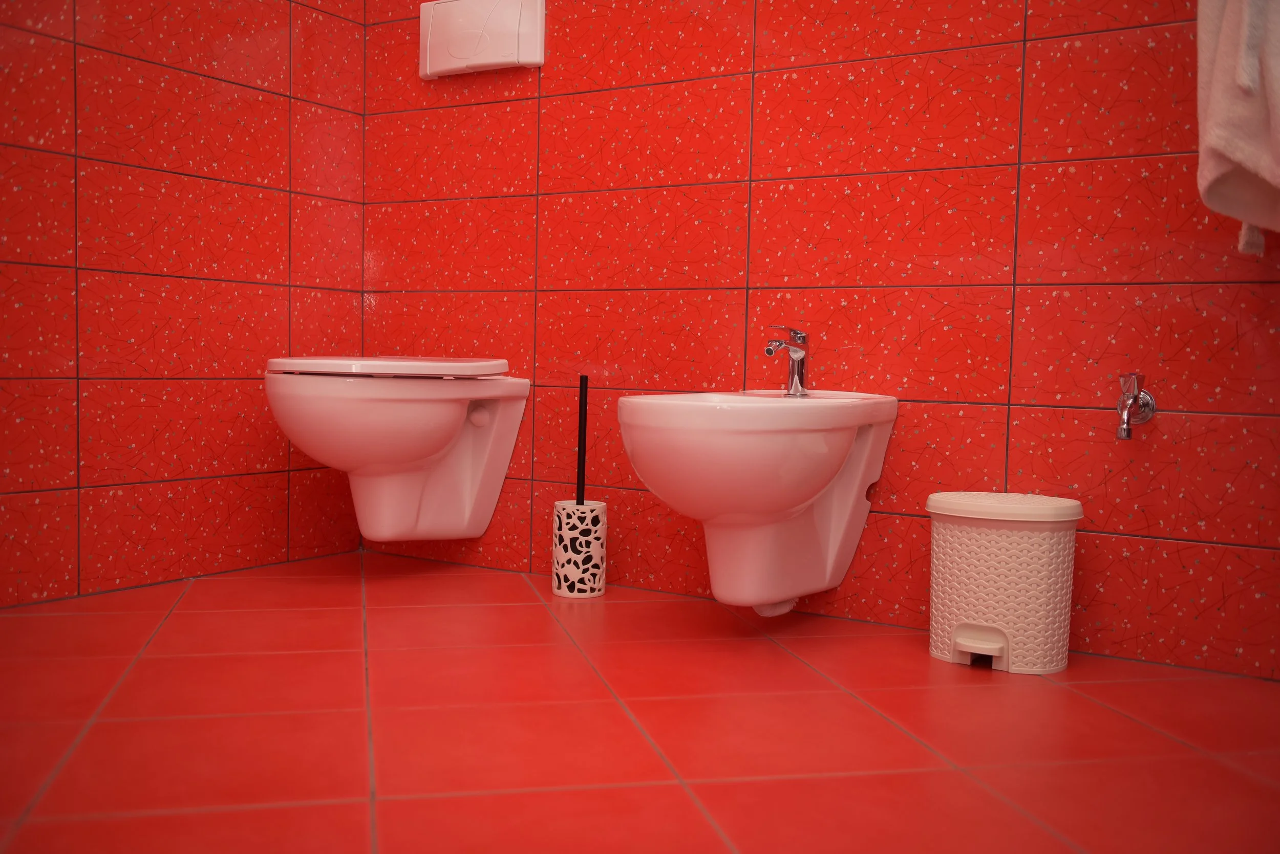Bagno con pareti e pavimento rivestiti con piastrelle rosse con decorazioni astratte. Vano toilette con water e bidet a parete, bicchiere porta spazzolini e cesto portaoggetti.