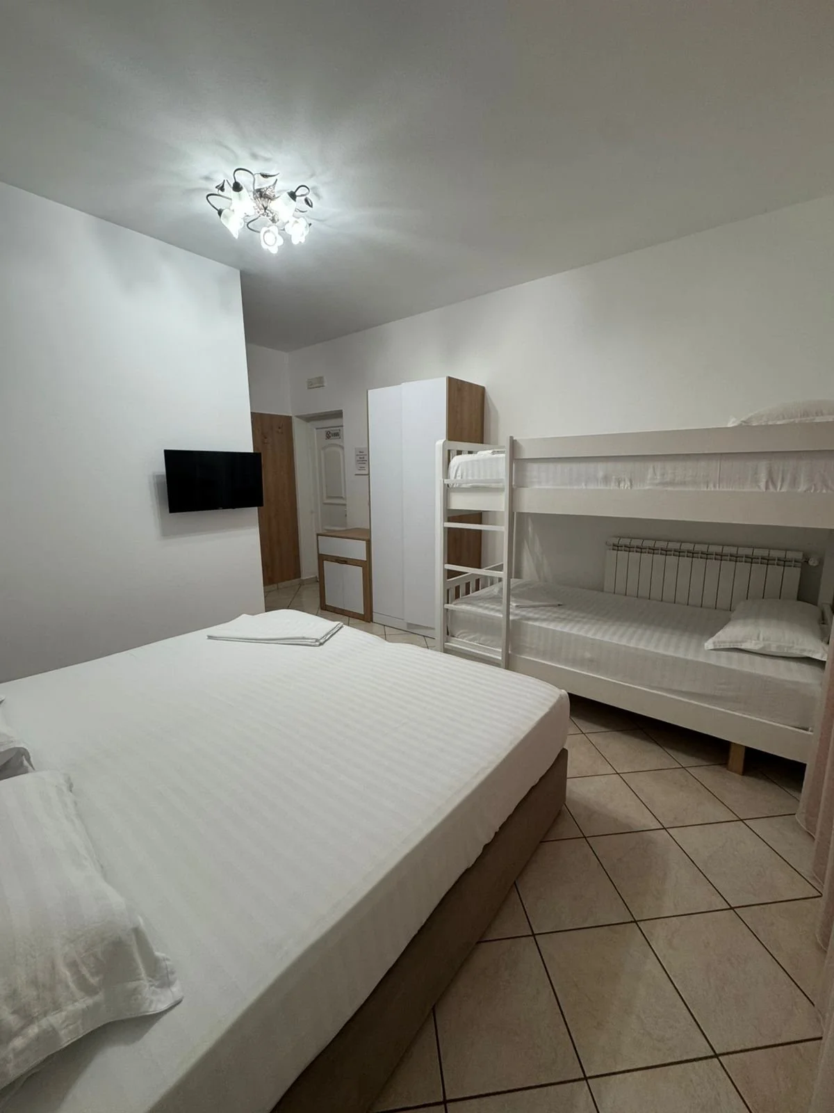 Camera con letto matrimoniale e letto a castello, televisore montato sulla parete, armadio bianco, soffitto con lampadario a più luci.