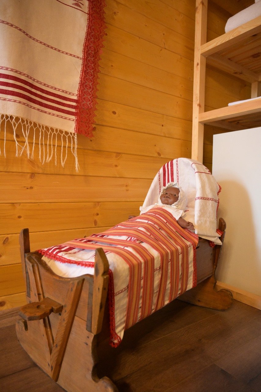 Statua di un bambino in culla di legno con coperta bianca e decorata con righe rosse. L'ambiente è in legno con pareti di assi di legno e cassettiere.