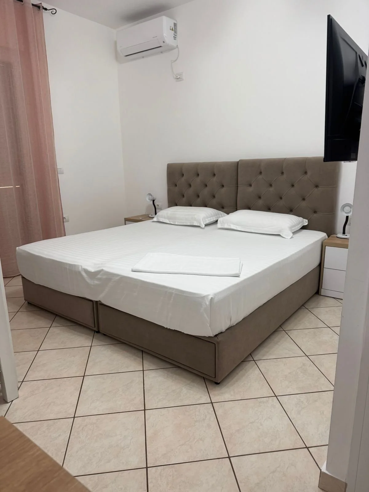 Letto matrimoniale con testiera imbottita color taupe e due cuscini bianchi, due comodini con lampade da lettura, aria condizionata e un televisore a parete in una stanza con pareti bianche e pavimento in piastrelle.