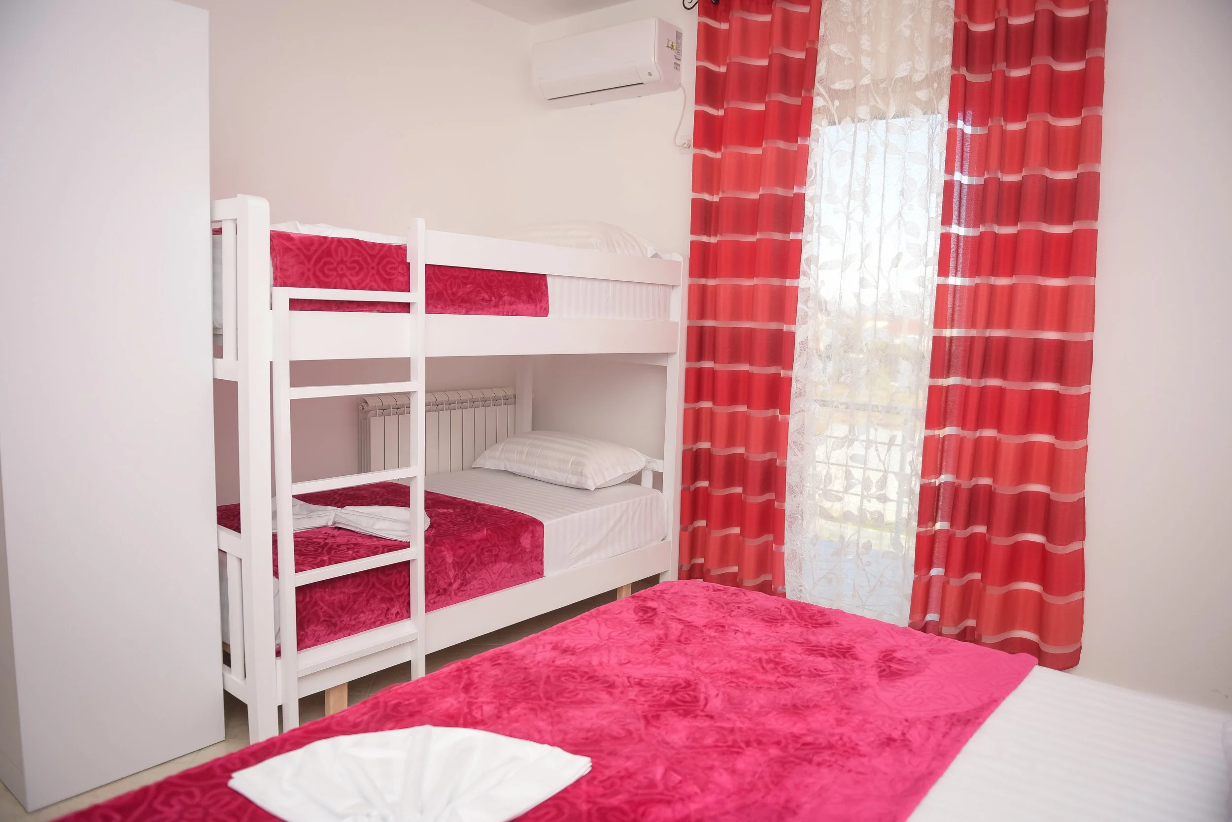 Camera con letto matrimoniale e letto a castello, tende rosse con boccini a righe, finestra con tenda trasparente e tende rosse, aria condizionata, pareti bianche.