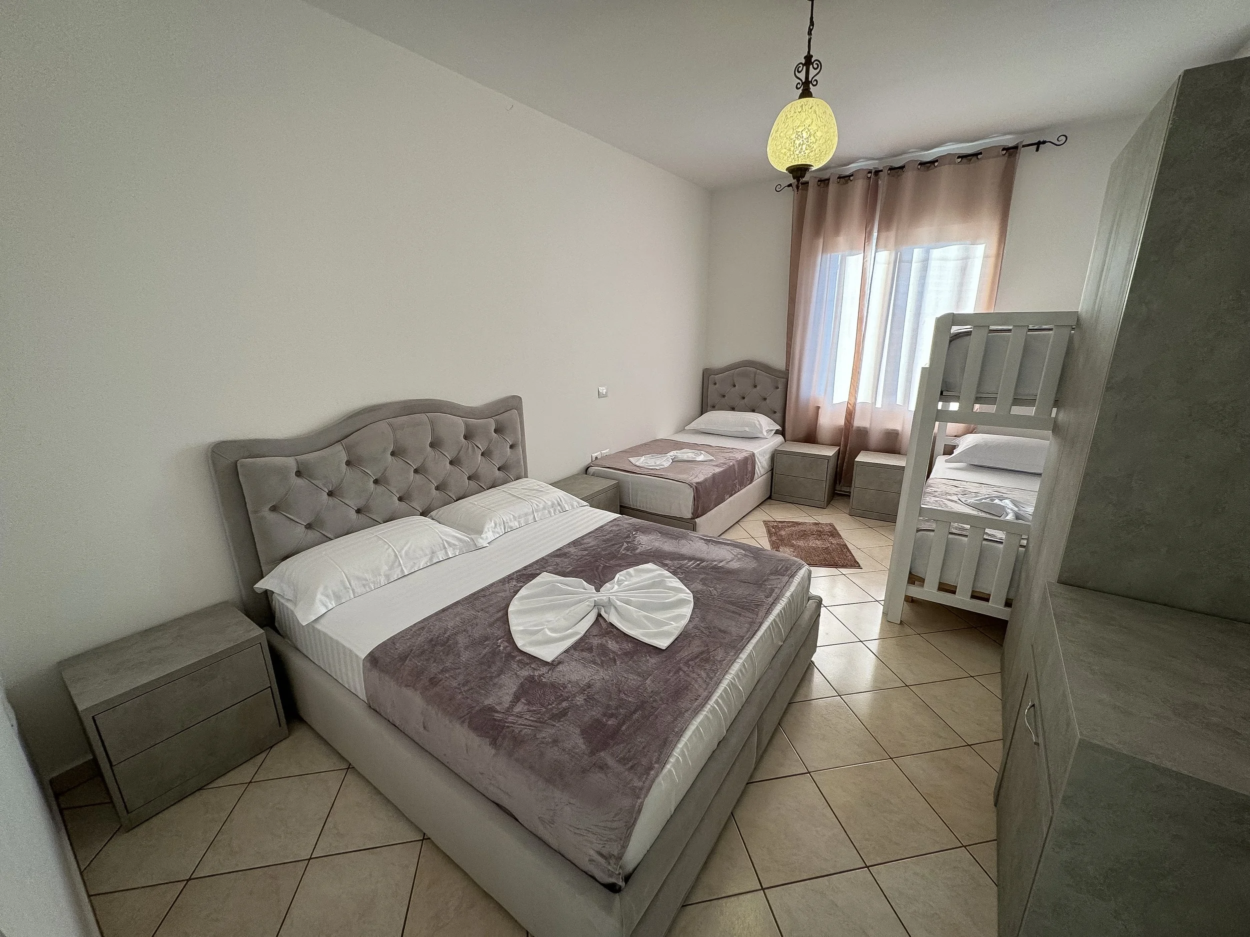 Stanza con due letti singoli, due comodini, grande finestra con tende rosa, lampadario e una cabina a castello.
