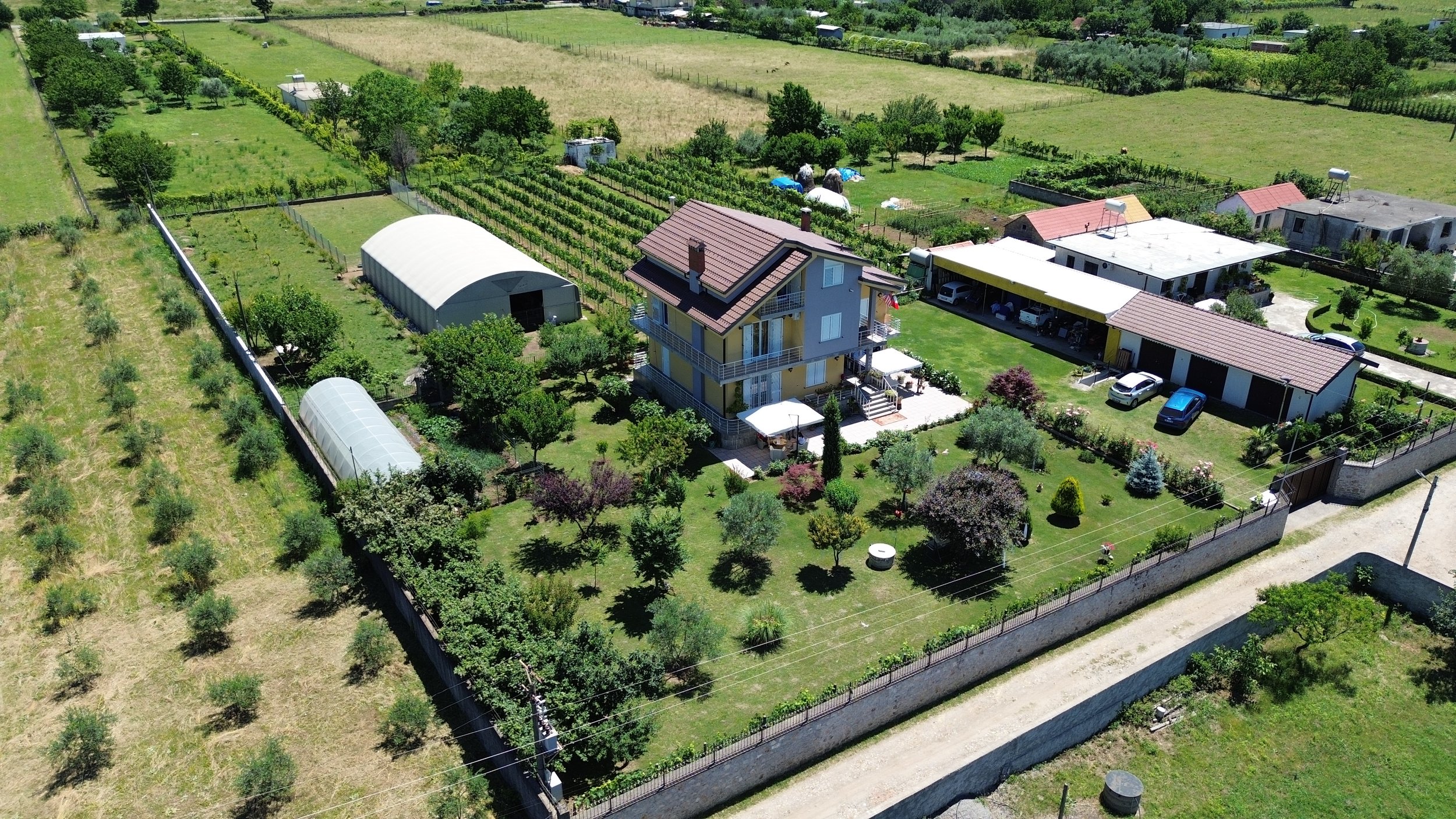 Un'ampia proprietà agricola con una casa di più piani circondata da un giardino ben curato, alberi e coltivazioni, con serre e strutture di supporto, il tutto incorniciato da campi di coltivazione e zone verdi.