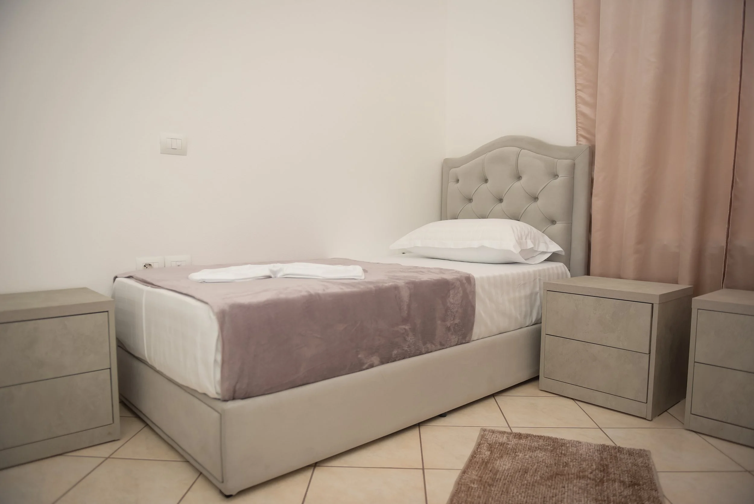 Letto matrimoniale con testiera imbottita beige, due comodini uguali alla destra e alla sinistra, due tappeti di cui uno di fronte al letto e uno di fianco, tende rosa chiaro, parete bianca con presa e interruttore.