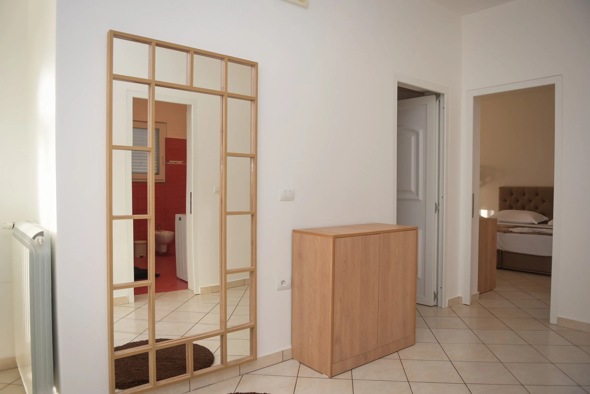 Interno di una casa con pareti bianche, una porta aperta su una stanza con un letto, e un divisorio di legno senza vetri.