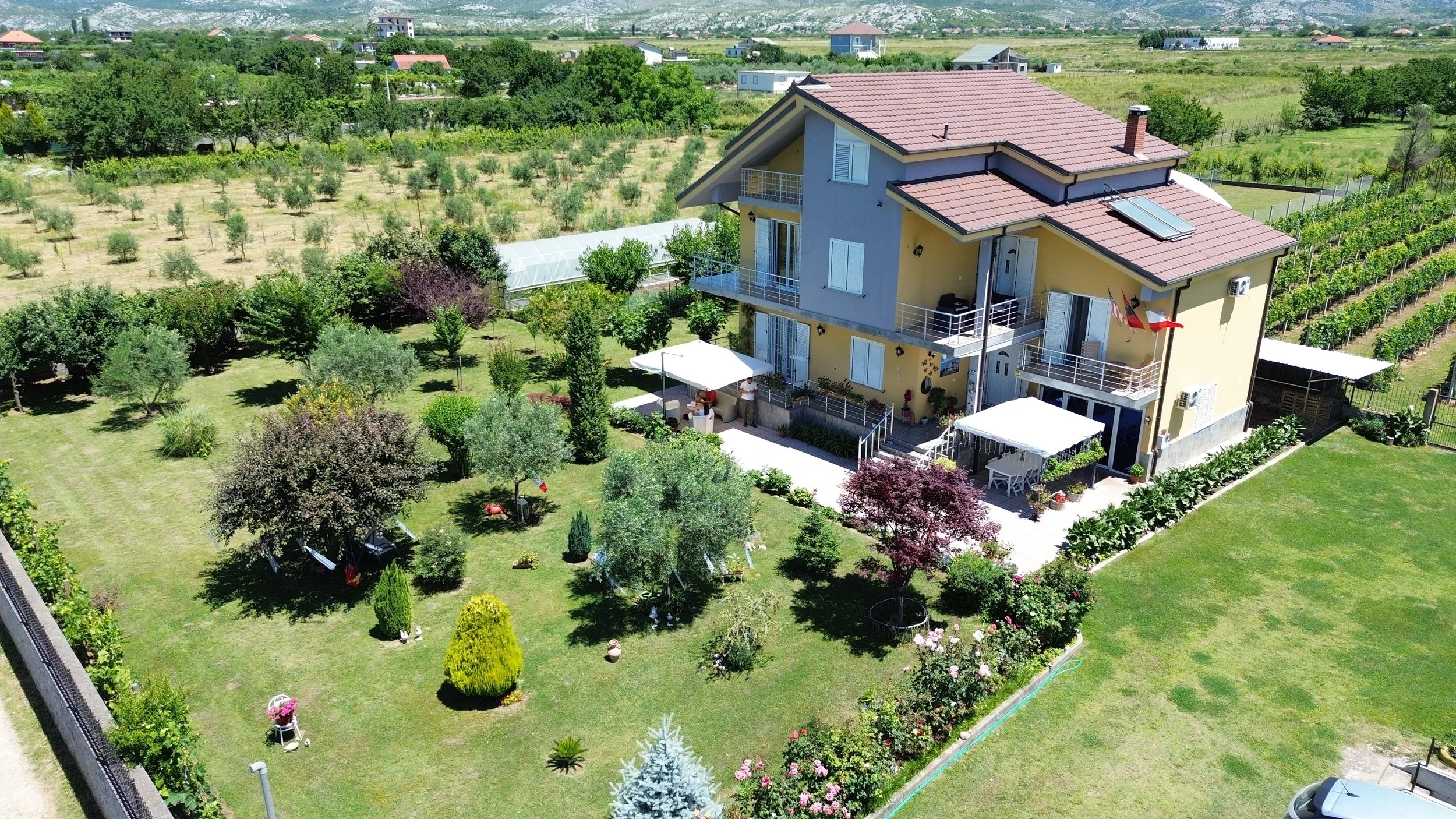 Casa moderna con balconi e giardino ben curato, circondata da alberi e piante, con campagna sullo sfondo.