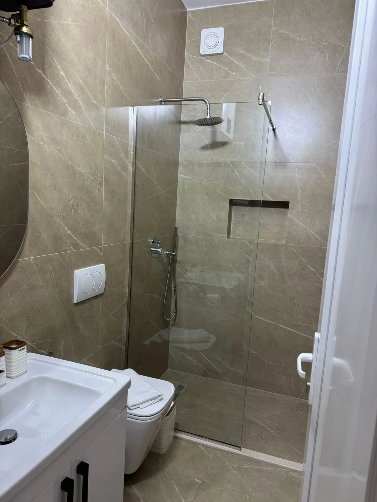 Bagno con pareti e pavimento in marmo beige, doccia con porta in vetro, soffione a pioggia, e accessori come specchio, porta asciugamani e vaso sanitario con bidet