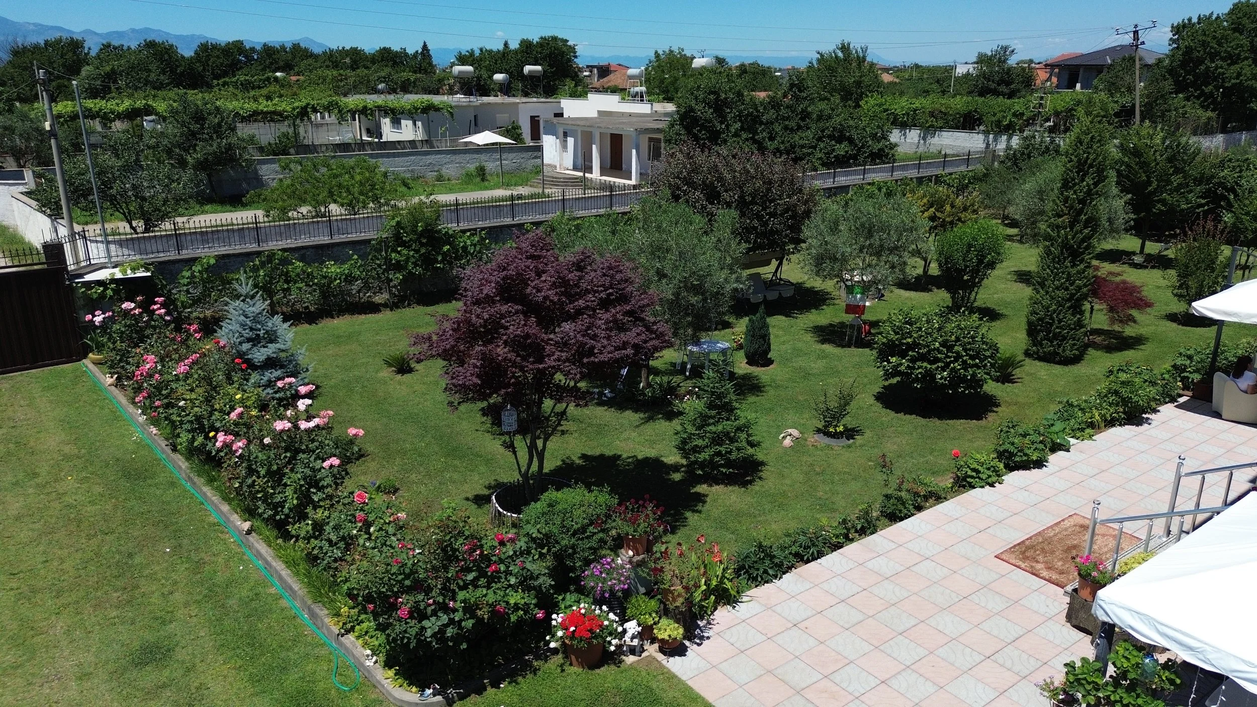 Giardino ben curato con vari alberi e fiori, spazio per sedersi con tavoli e ombrelloni, recinzione e strade circostanti, in un'area residenziale con montagne sullo sfondo.