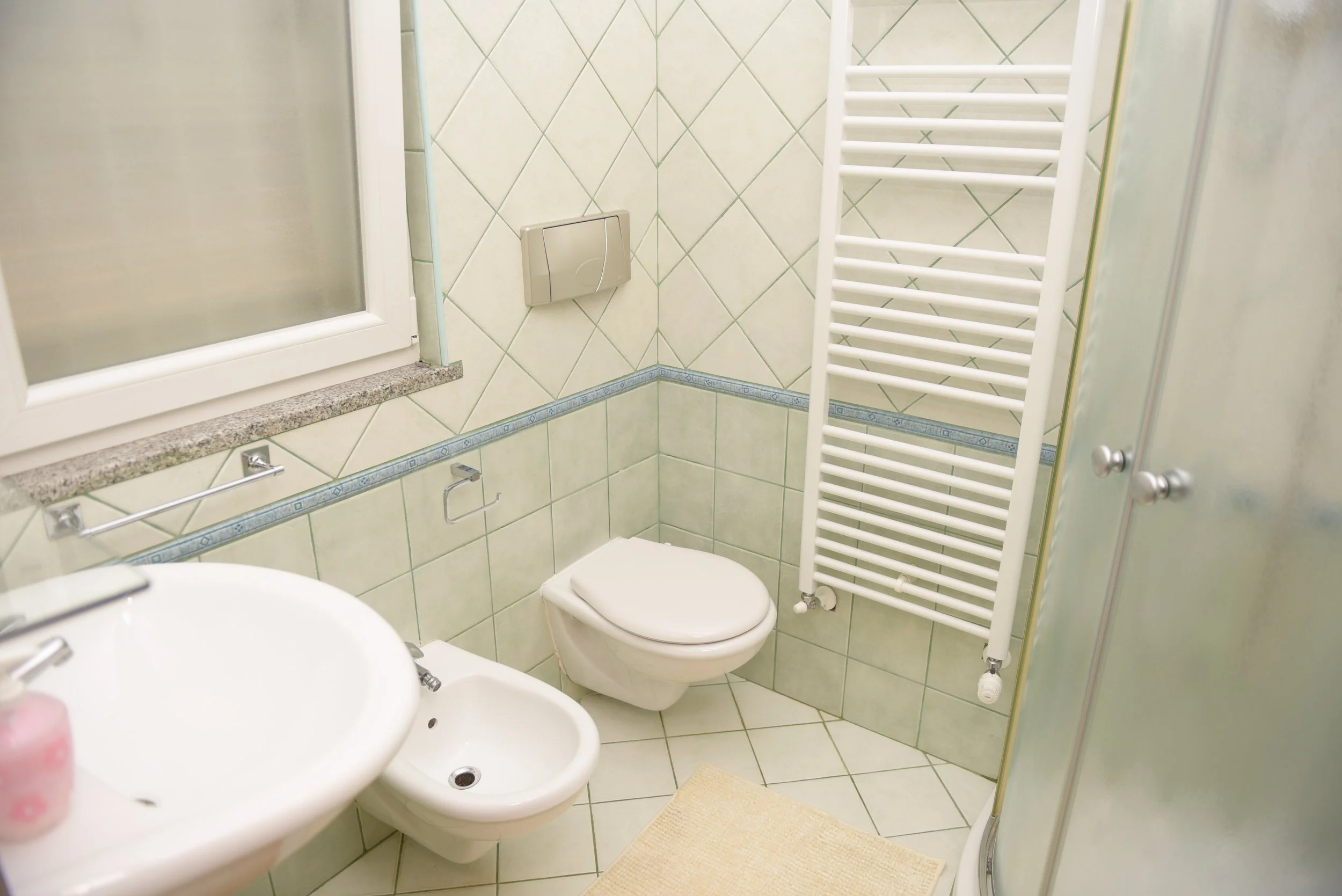 Bagno con WC, bidet, lavabo, radiatore bianco, finestra con cornice bianca e pareti piastrellate in beige e azzurro.