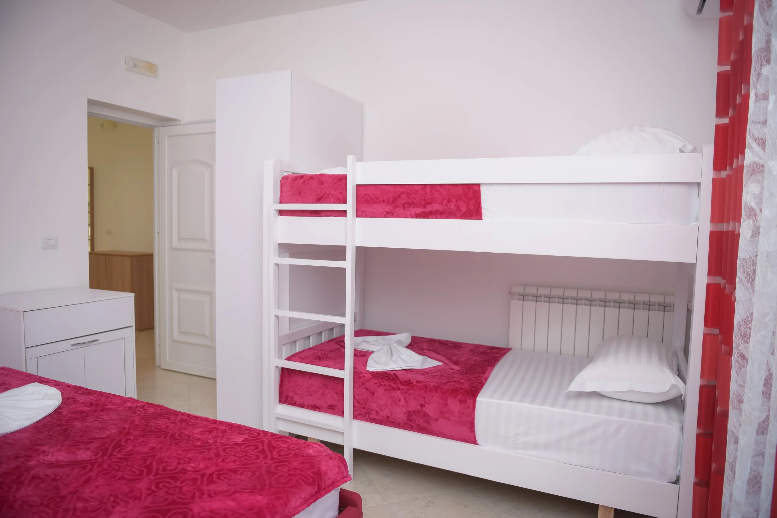 Camera con un letto a castello bianco, con lenzuola rosse e cuscini, in una stanza con pareti bianche e tende rosse e bianche, e una porta bianca aperta sulla sinistra.
