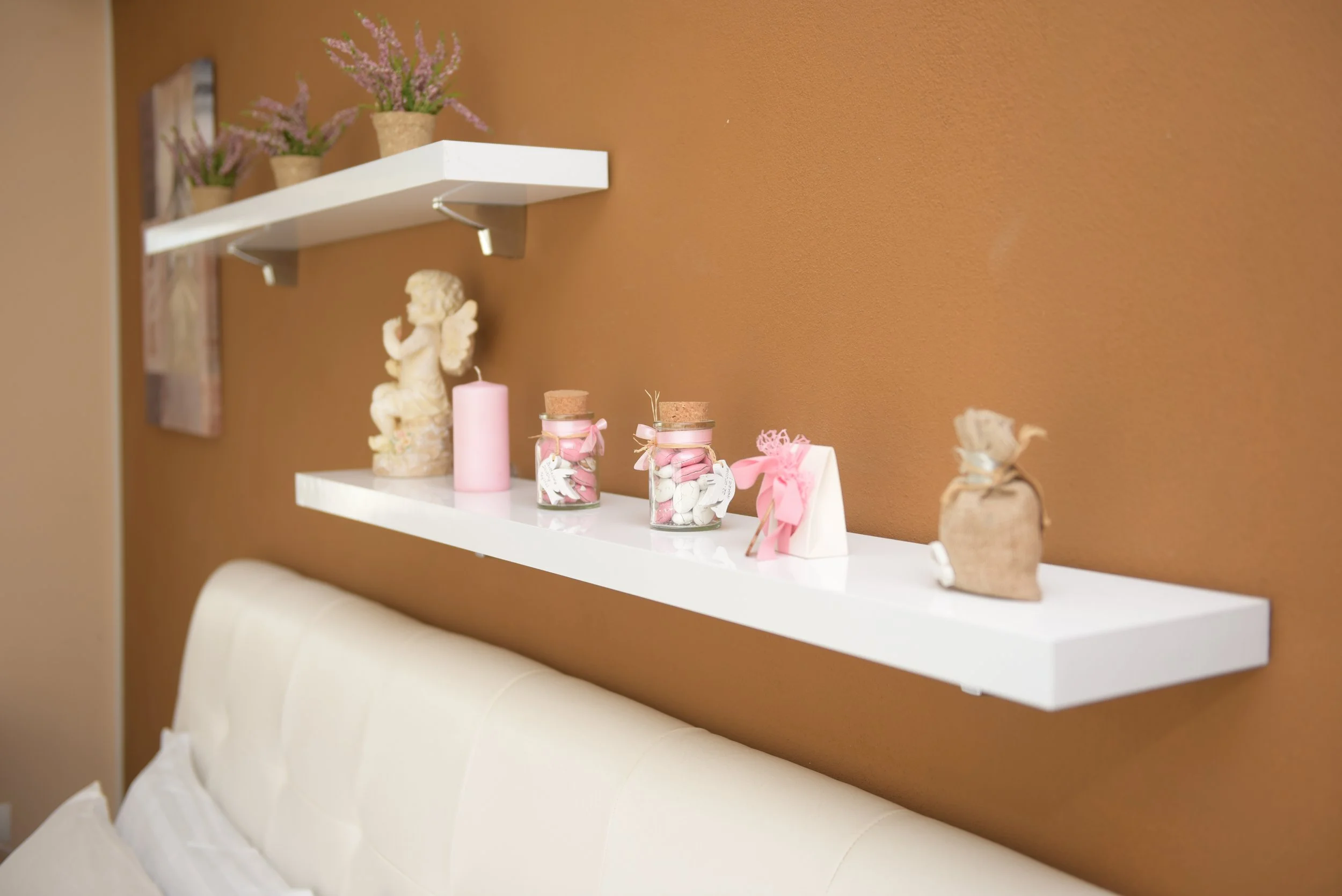 Mensola bianca con decorazioni, tra cui vasi di fiori, candele rosa, barattoli con decorazioni e un angelo di pietra. Sfondo di parete marrone.