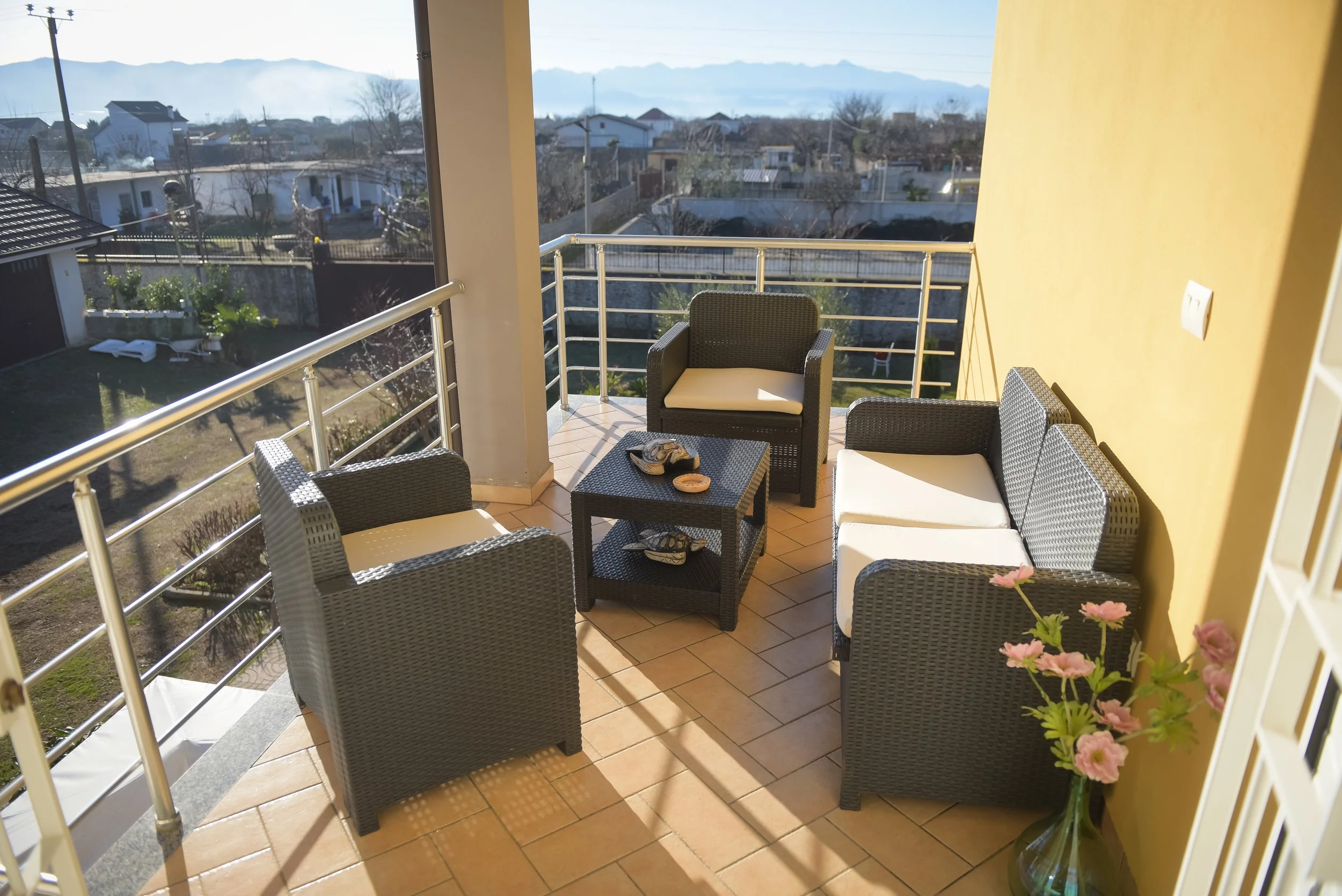 Balcone arredato con sedie e tavolino, fiori rosa in vaso, vista su cortile e case, montagne sullo sfondo.